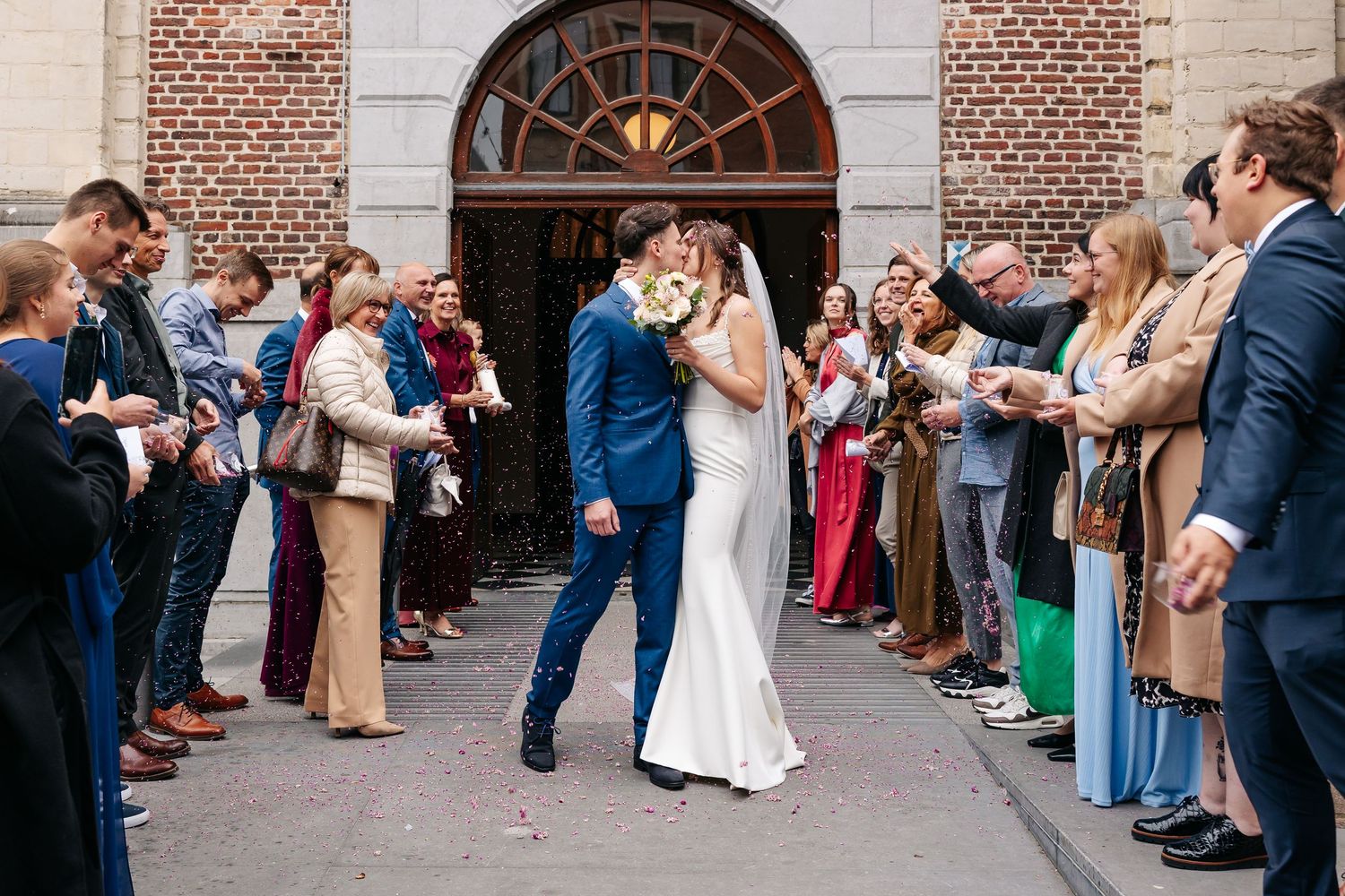Newlyweds share a romantic kiss outside church surrounded by cheering wedding guests.