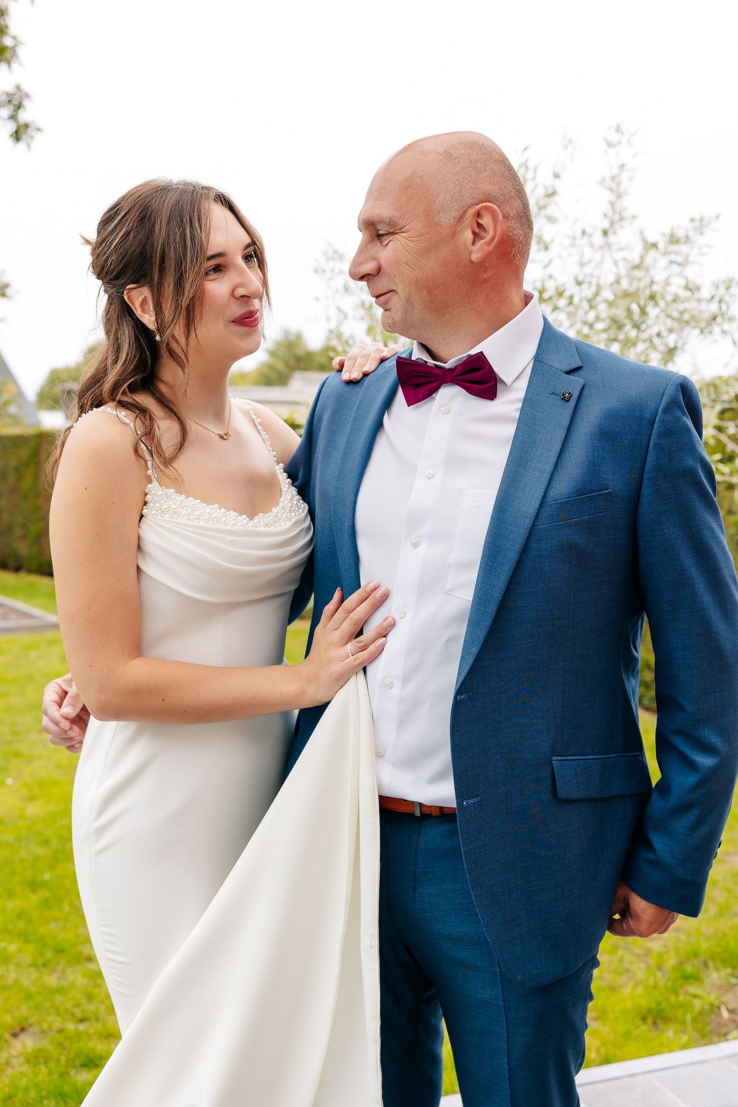 Bride in white gown and groom in blue suit with burgundy bow tie share a tender moment outdoors.