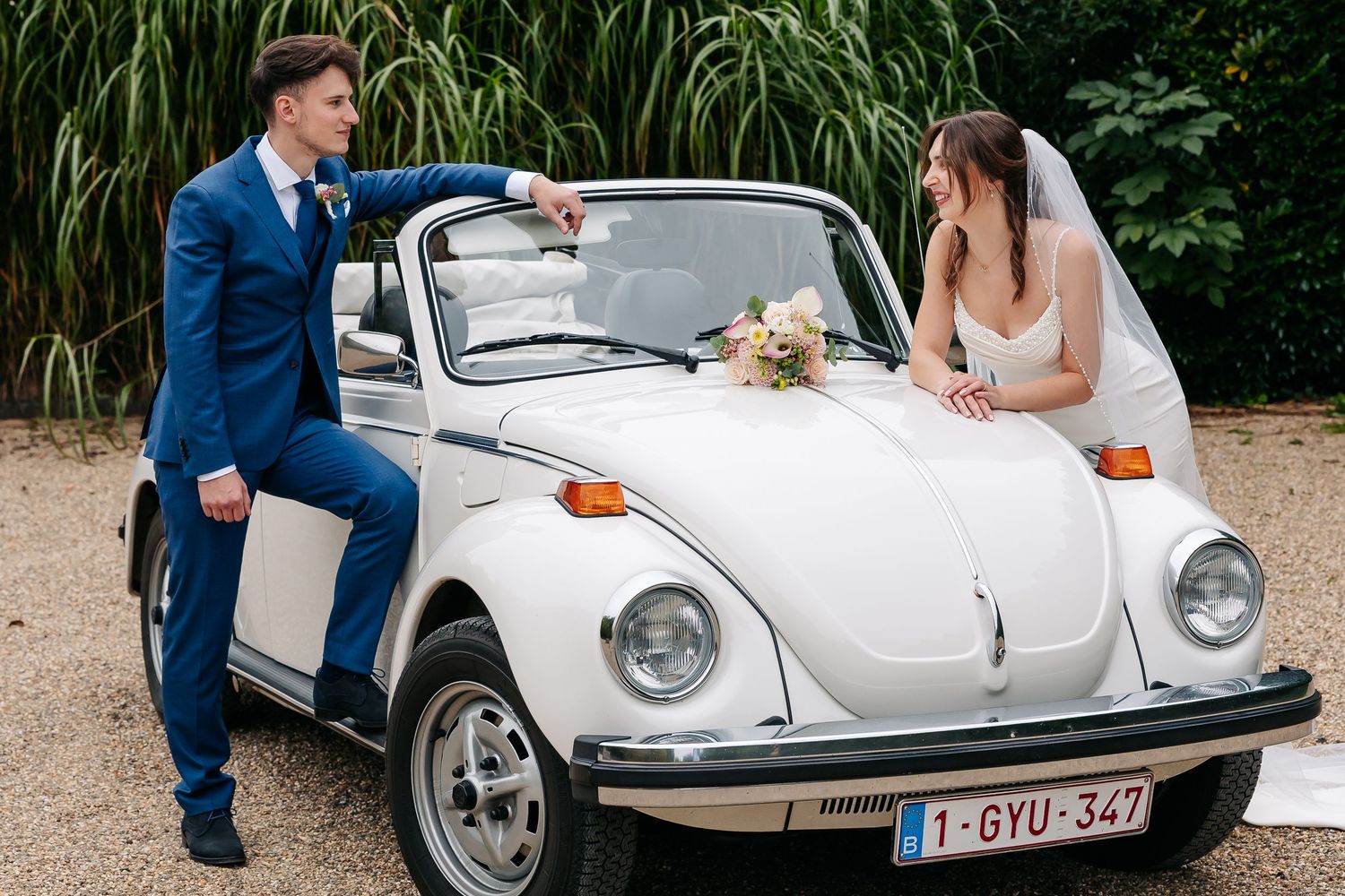 Bride in white gown and groom in blue suit posing with a classic white VW Beetle convertible.