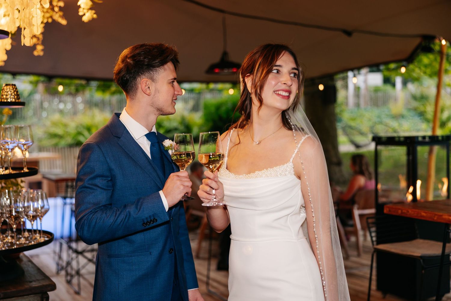 Bride and groom clinking white wine glasses and smiling under a decorated wedding reception canopy.