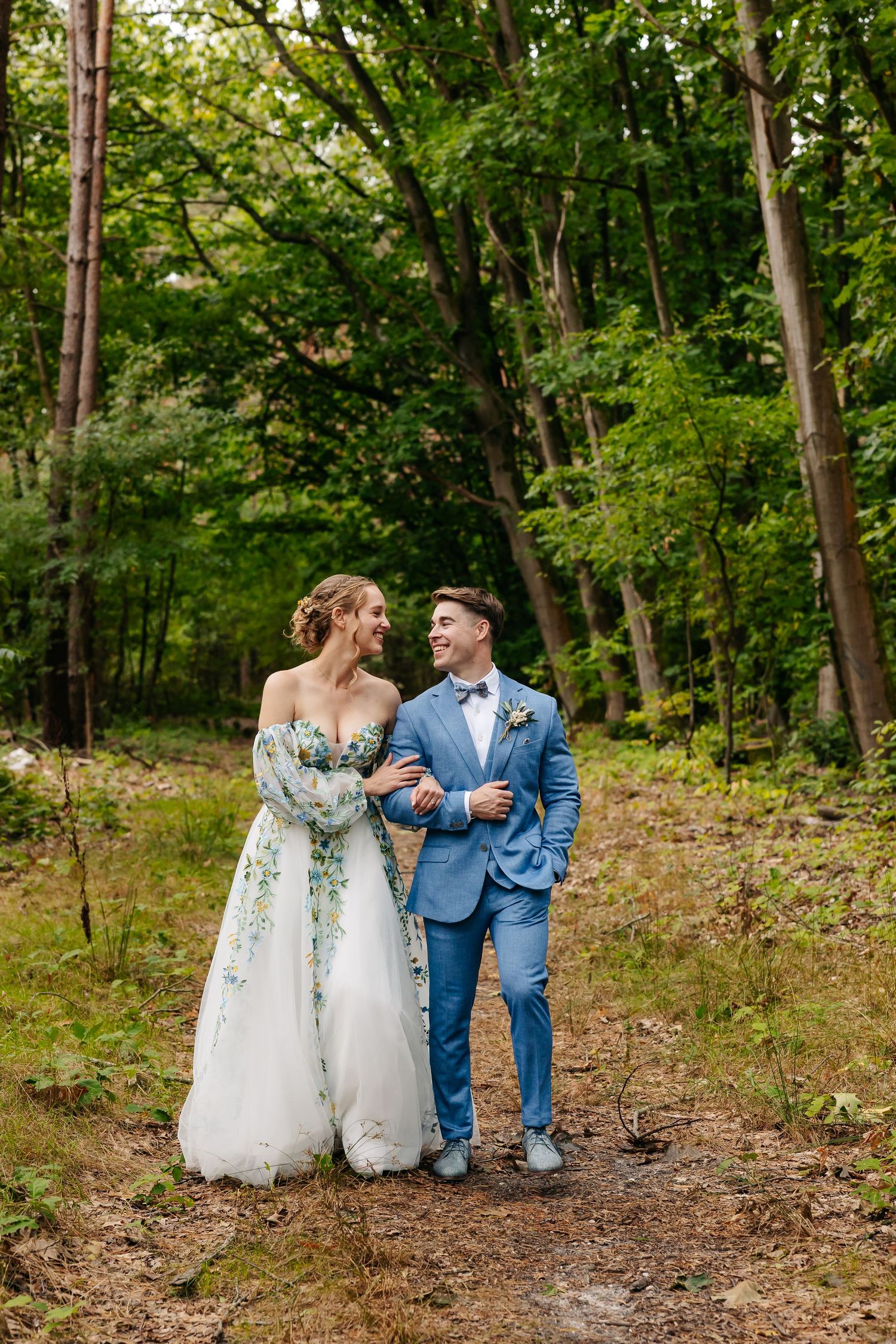 Bride in floral gown and groom in blue suit walking in forest.