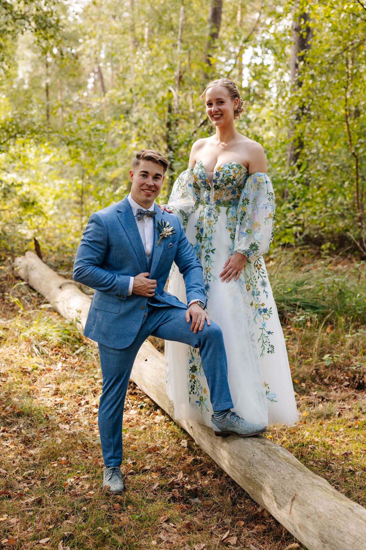 A bride in a floral gown and groom in a blue suit pose together on a fallen log in a forest.