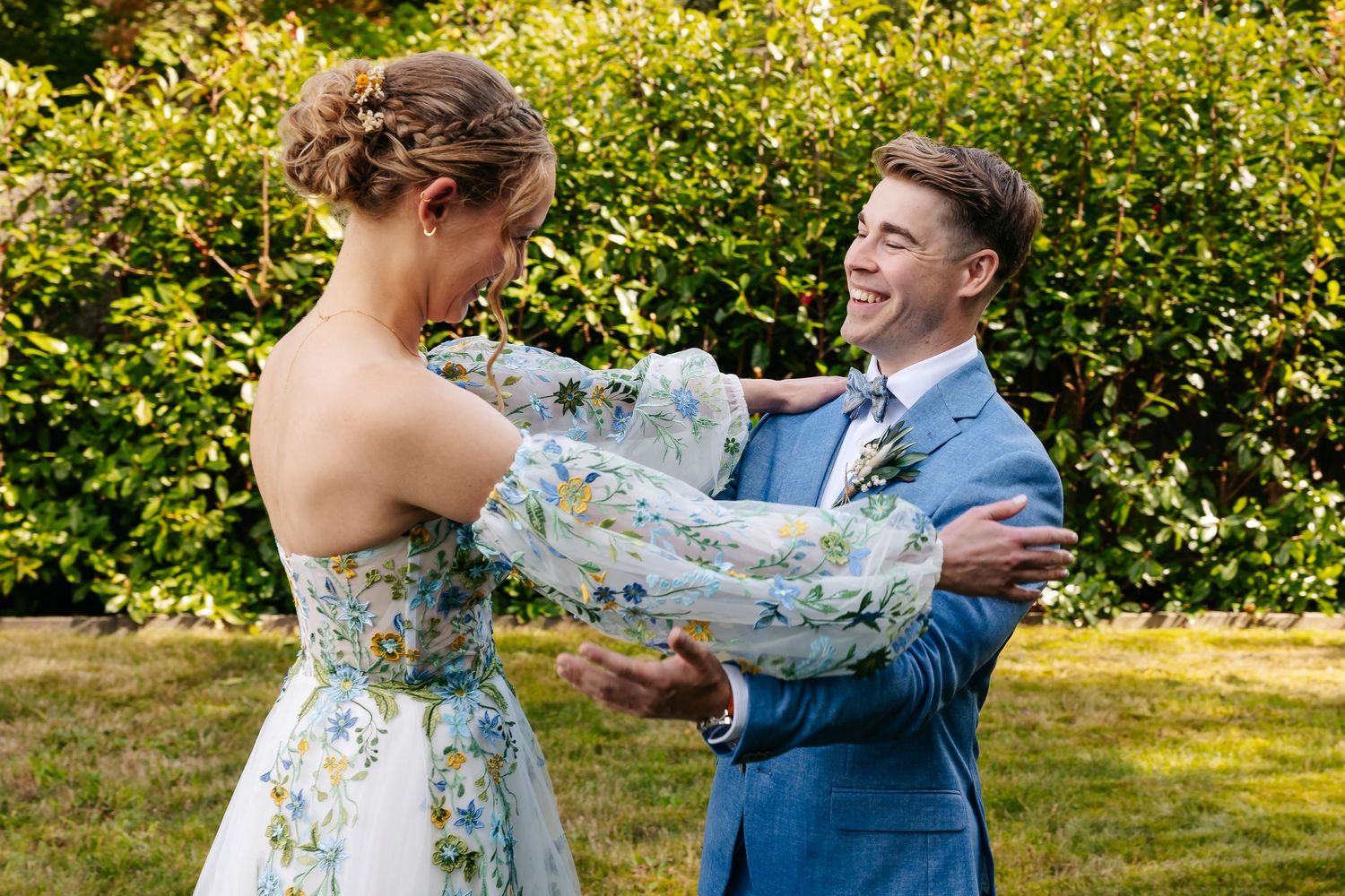 Groom in blue suit laughs joyfully as bride in floral dress reaches out to him in garden setting.