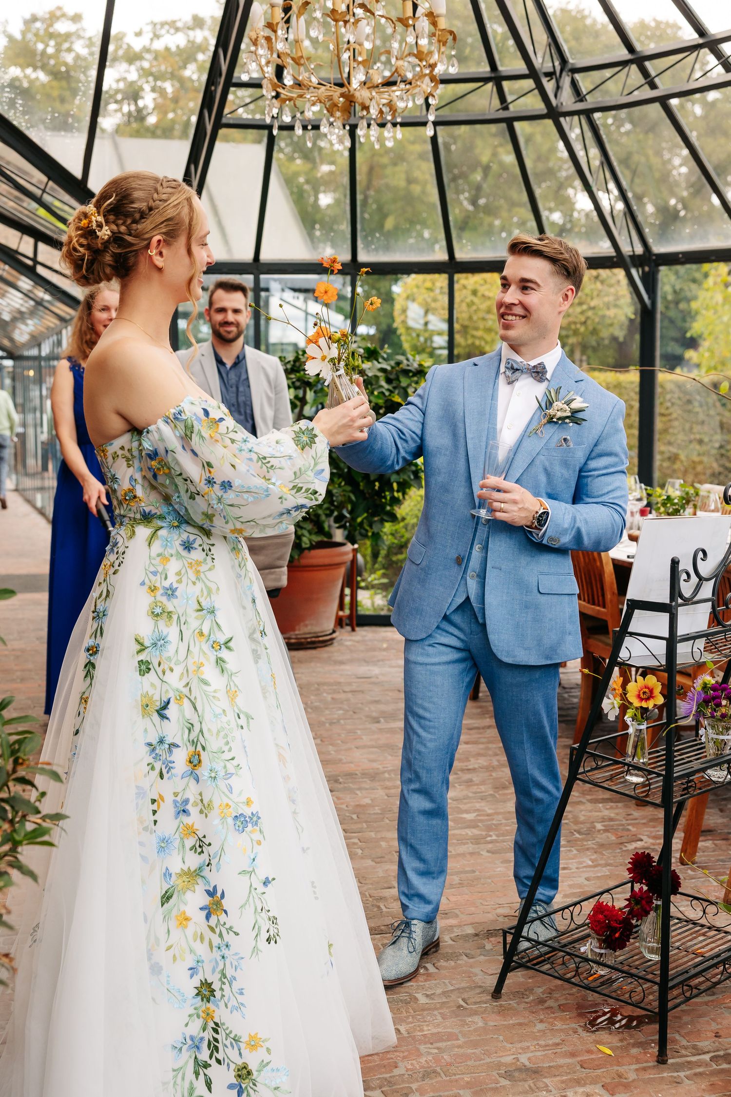 Bride in floral gown and groom in blue suit sharing a joyful moment inside glass greenhouse.