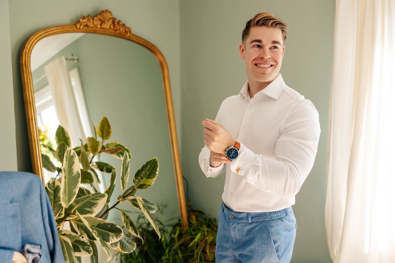 Smiling groom in white shirt adjusting watch while standing in front of ornate gold mirror.