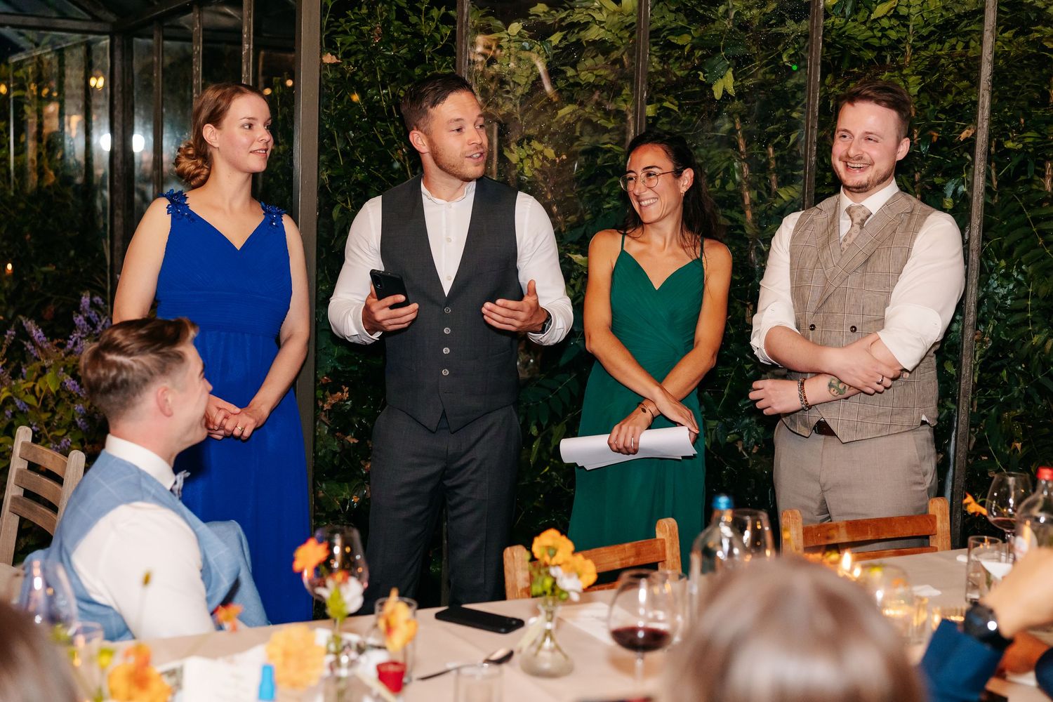 Four elegantly dressed wedding guests stand and smile during a reception speech at a candlelit dinner.