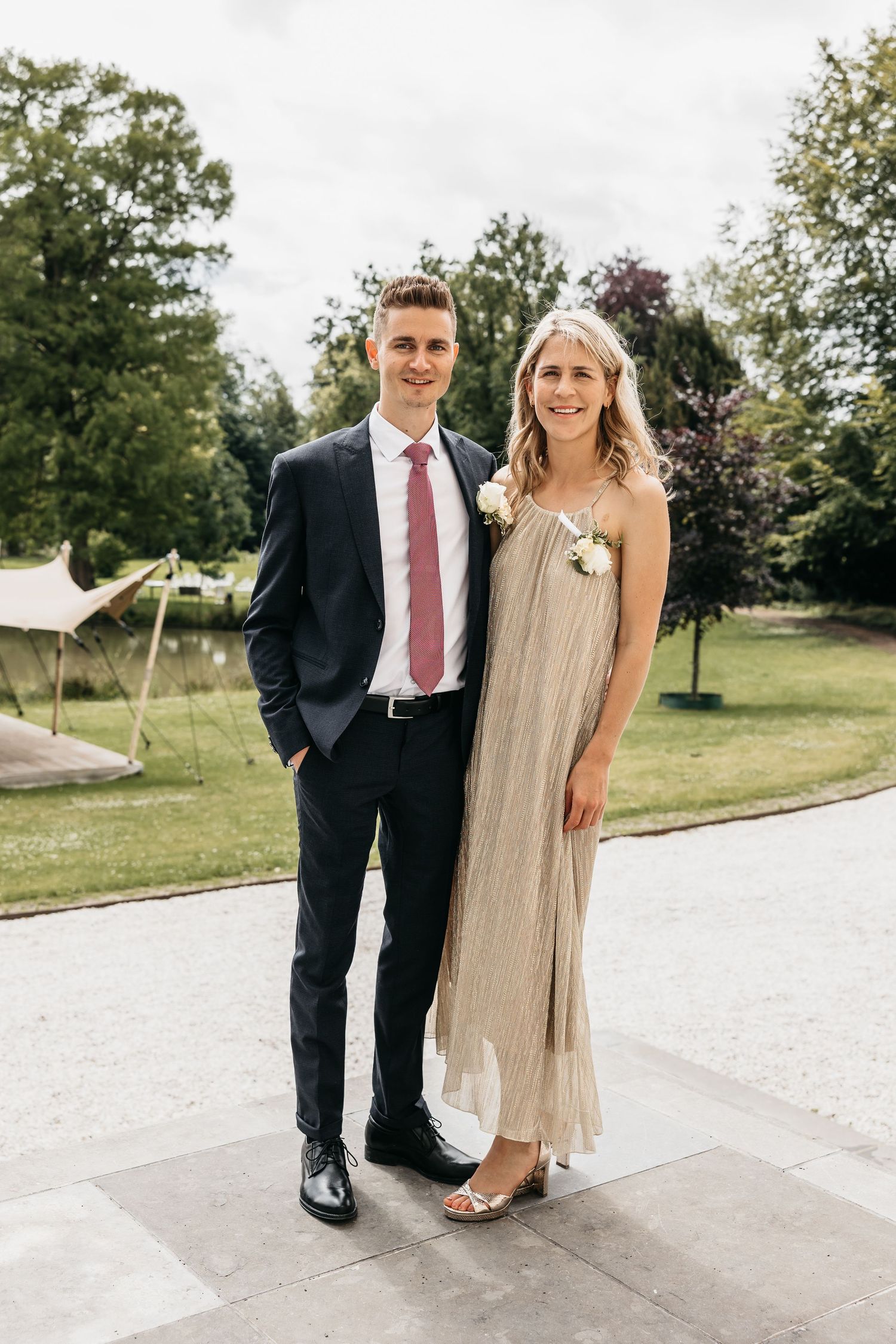 Elegant wedding guests pose outdoors, man in navy suit with pink tie and woman in gold pleated dress with floral corsage.