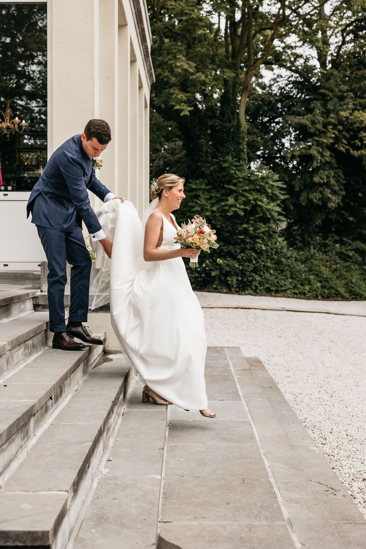 Groom helps bride carry her white wedding gown train down outdoor stone steps.