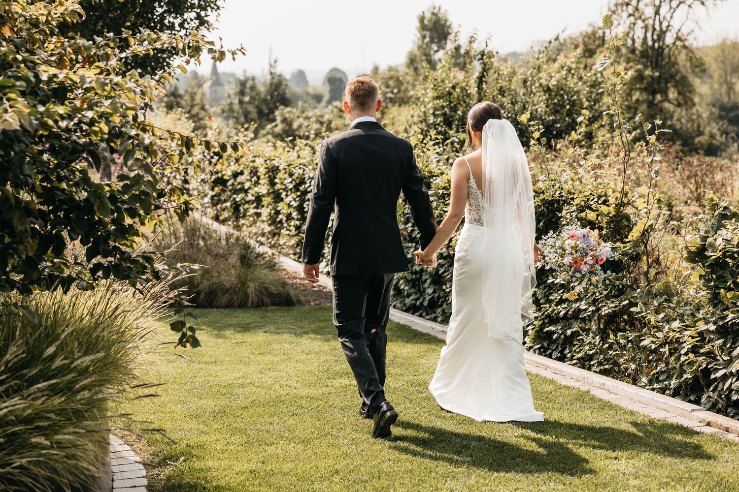 Bride and groom walking away hand in hand through a lush garden pathway on their wedding day.