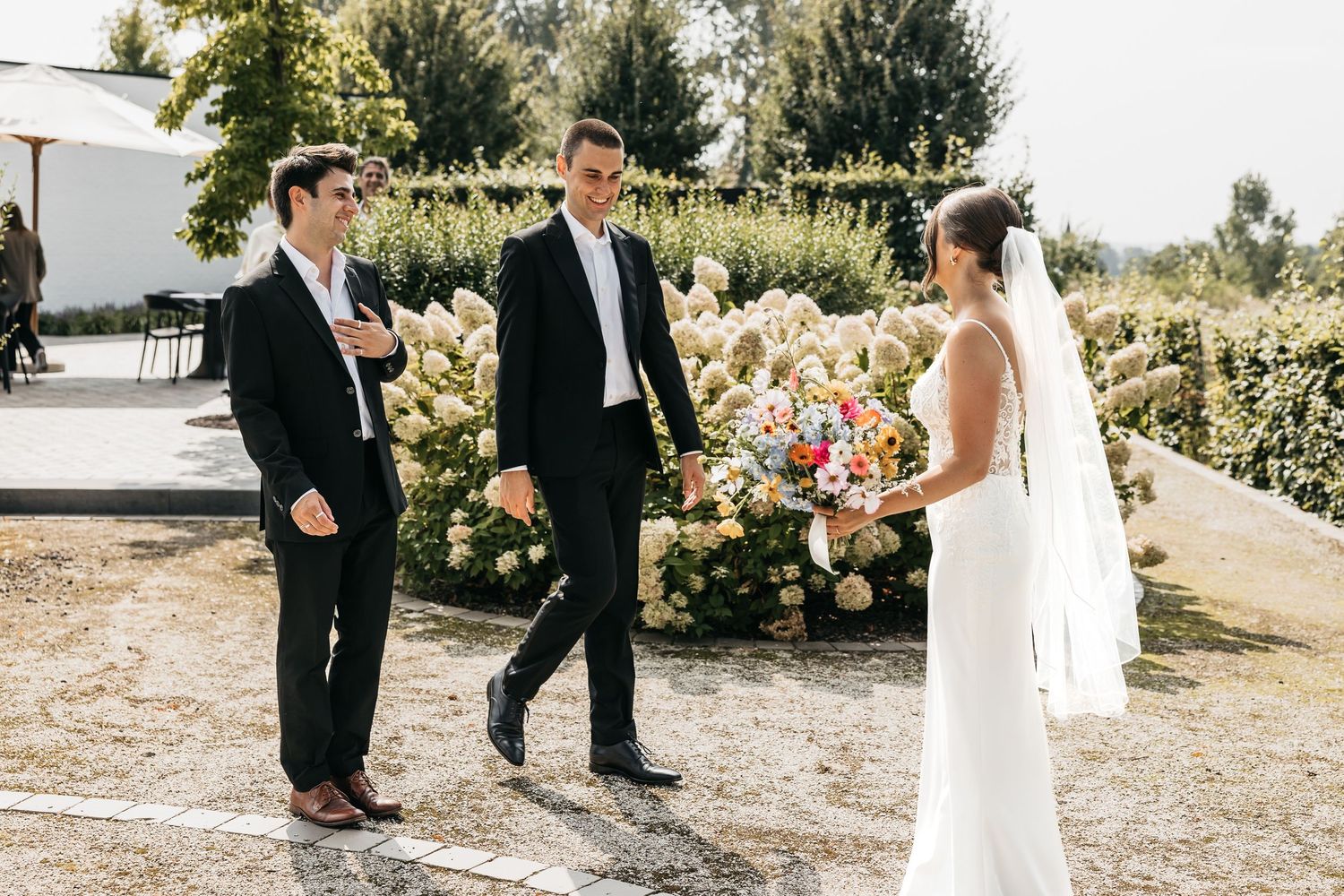 Bride in white gown holding colorful bouquet greeting two groomsmen in black suits in sunny garden.