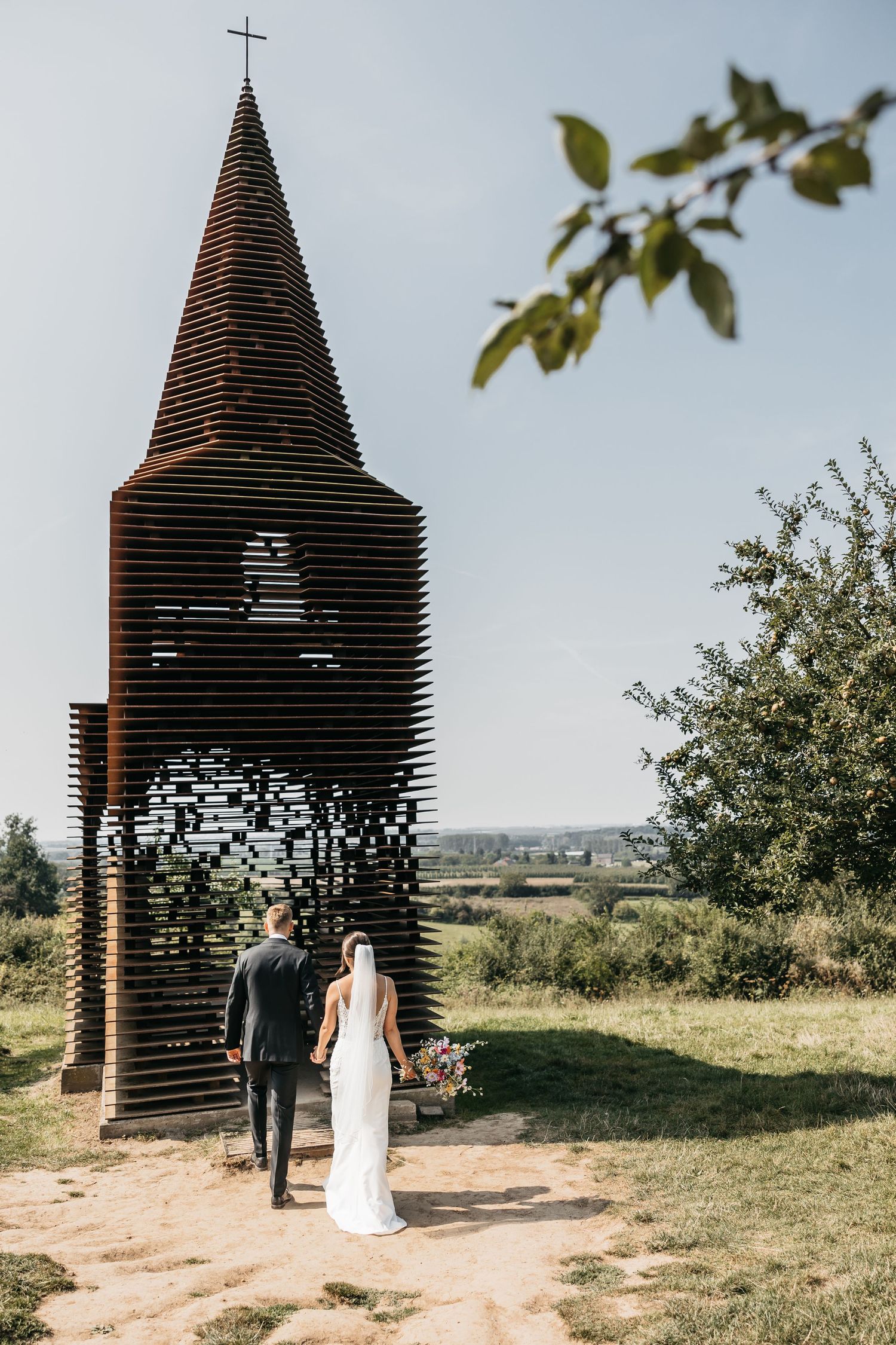Bride and groom walk hand in hand toward a stunning modern wooden chapel tower under a clear blue sky.