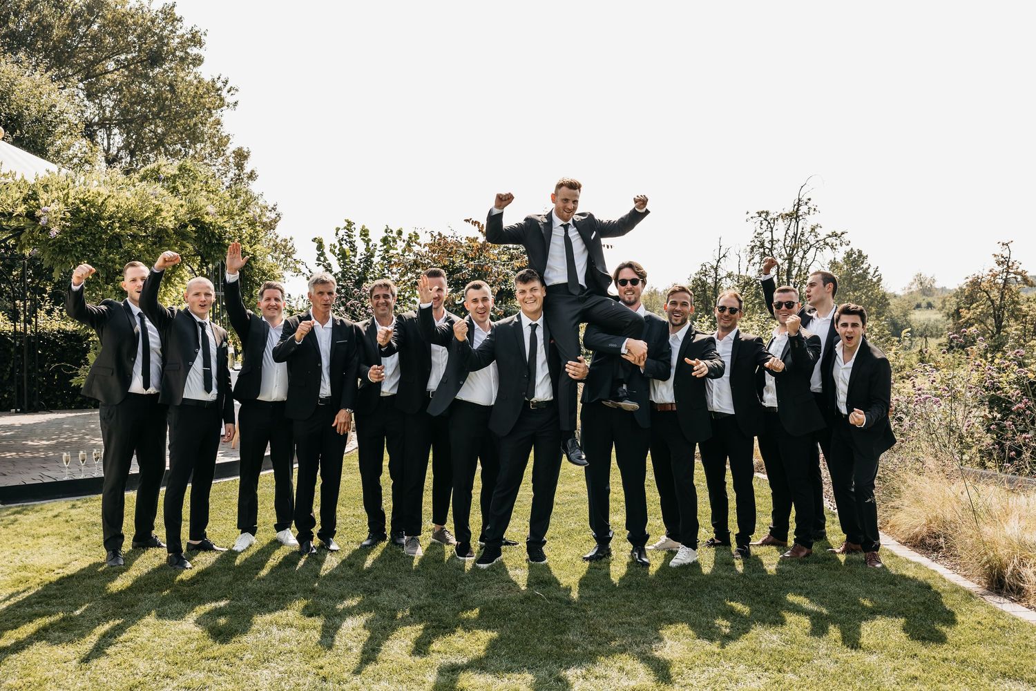 Large groomsmen group cheering as groom is lifted in the air at outdoor wedding.