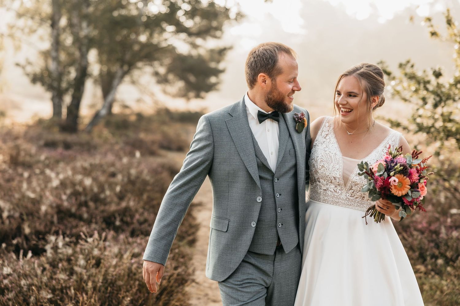 Bride and groom walking closely together through a sunlit heathland, sharing a joyful smile.