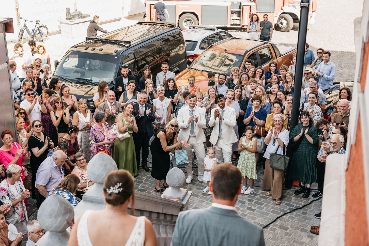 Aerial view of large wedding crowd cheering for bride and groom on steps outside venue with parked vehicles.