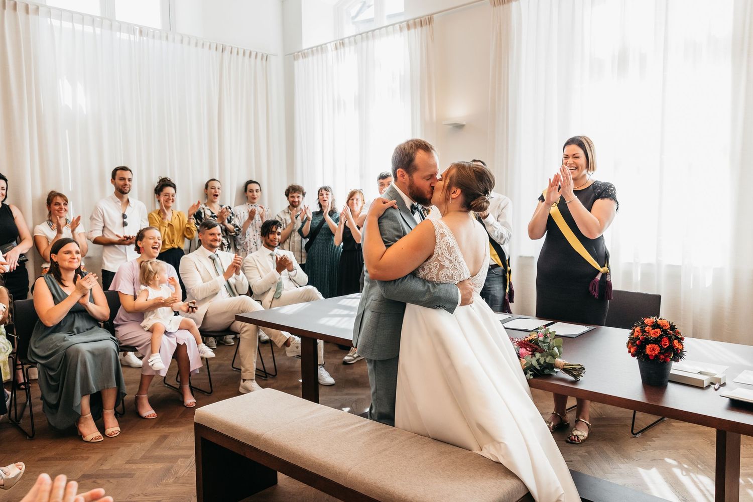 Newlyweds share their first kiss at a civil wedding ceremony while guests applaud in a bright elegant room.