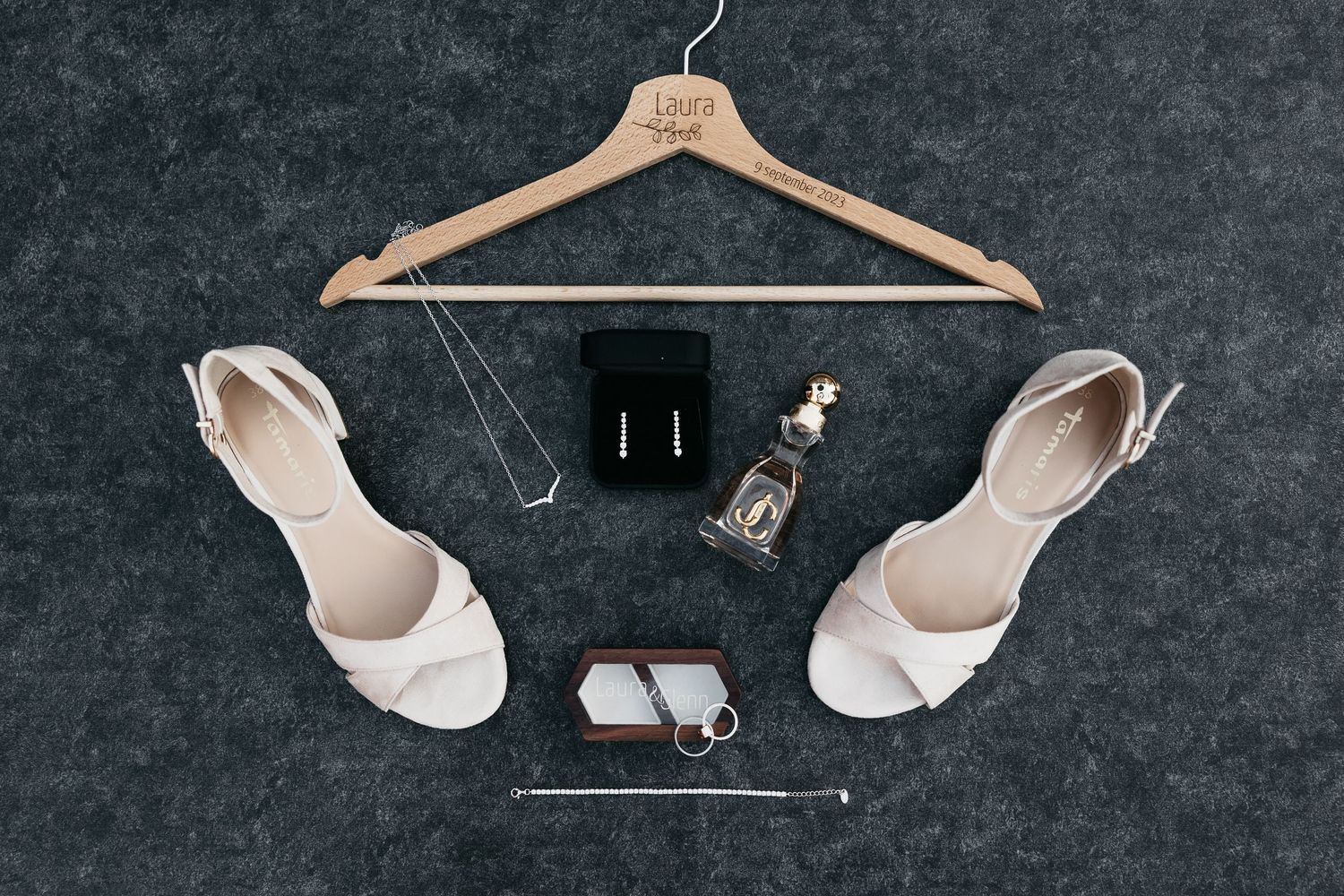 Flat lay of bridal accessories including white heels, earrings, perfume, and personalized hanger.