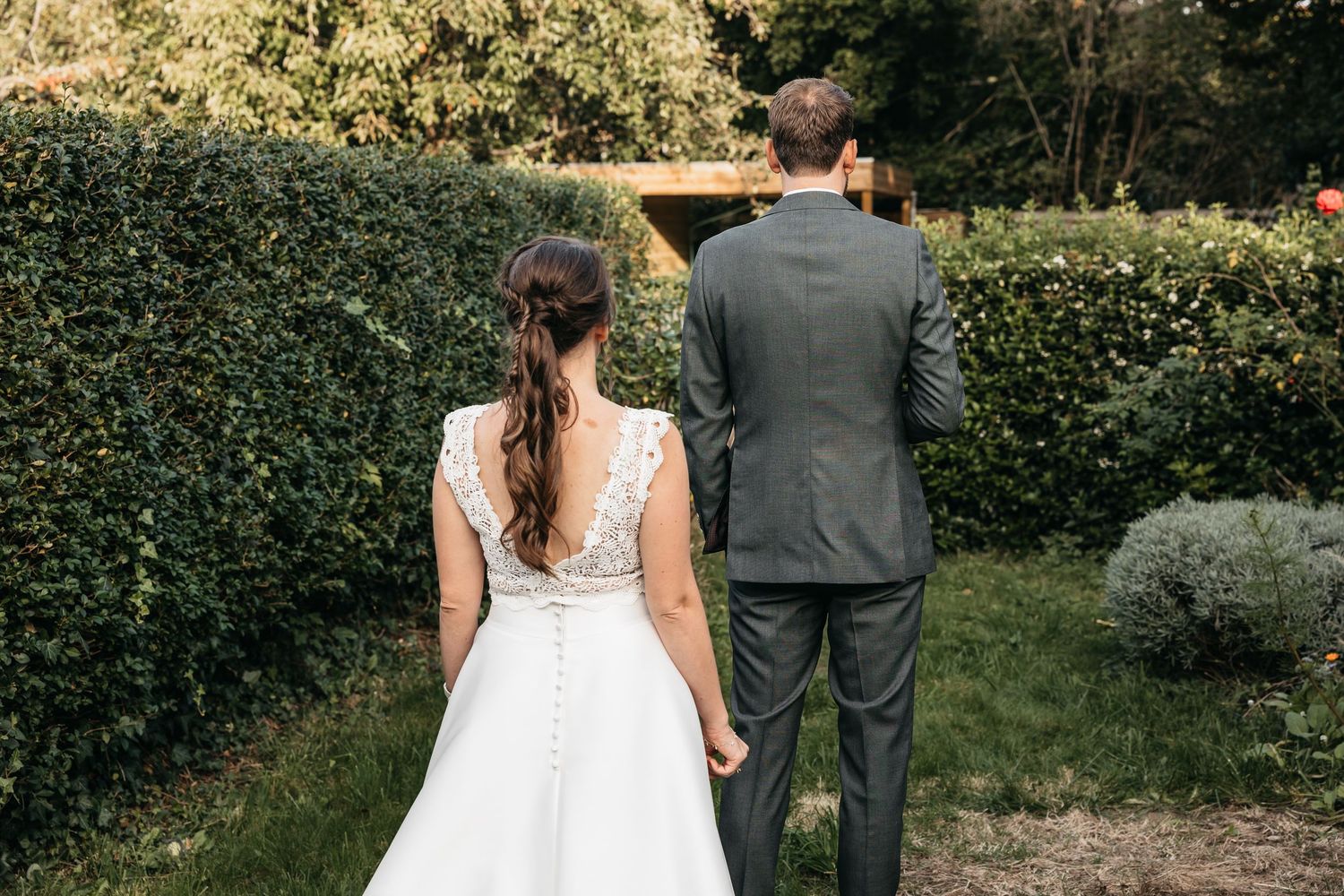 Wedding couple walking hand in hand through lush green garden, bride in white lace gown and groom in gray suit.
