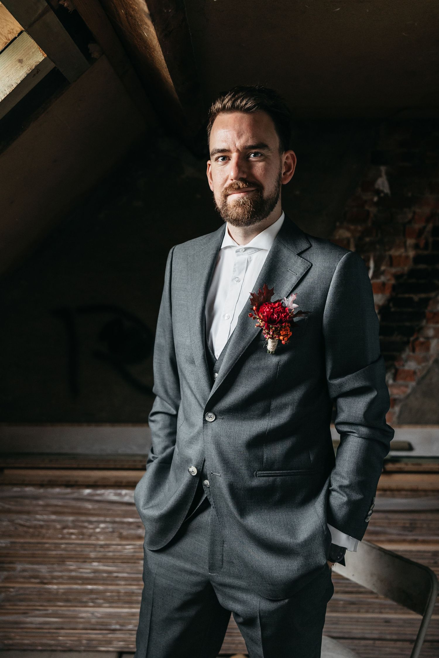Confident groom in gray suit with red boutonniere posing hands in pockets in rustic loft.