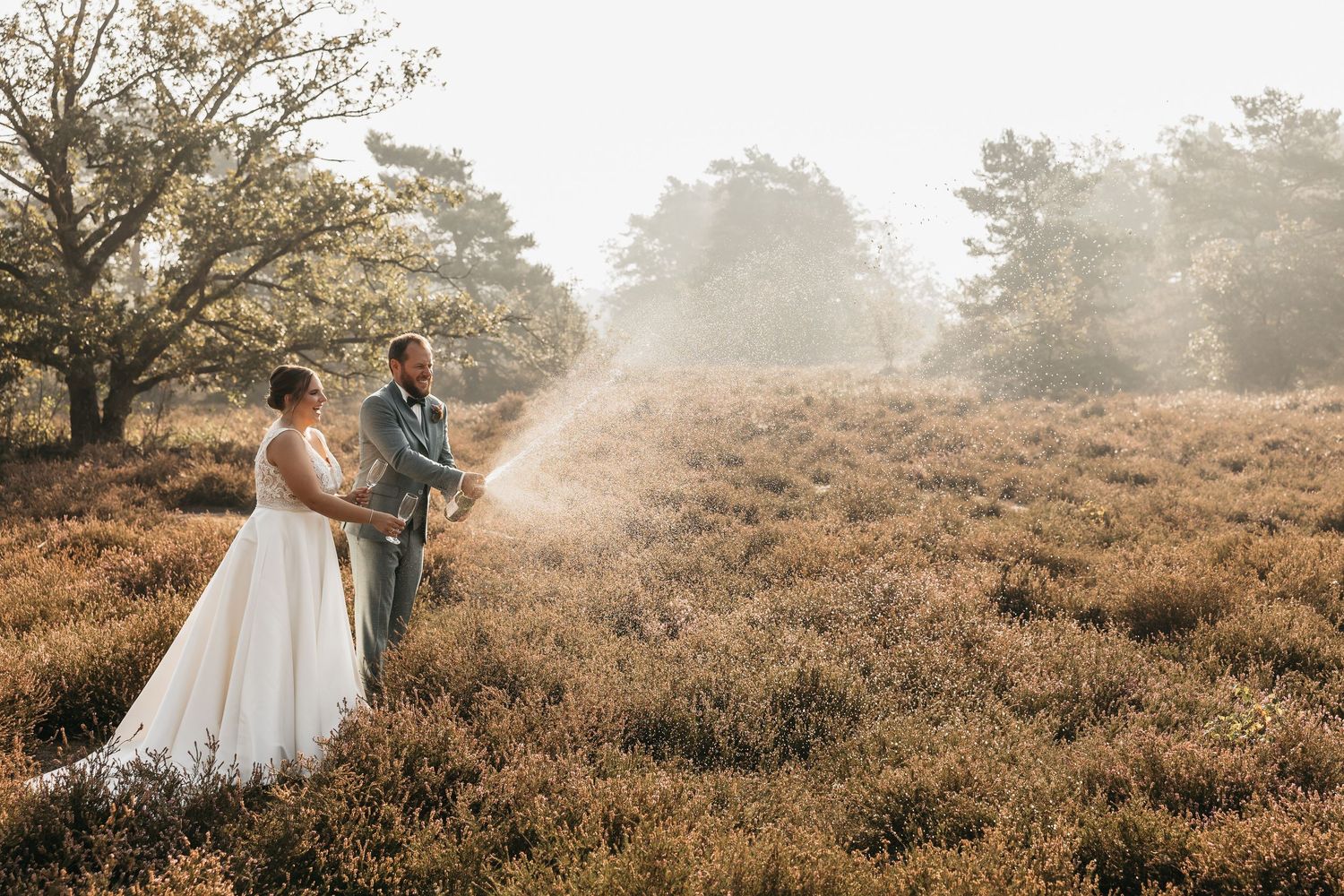 Bride and groom standing together as champagne sprays in misty golden heathland wedding photo.