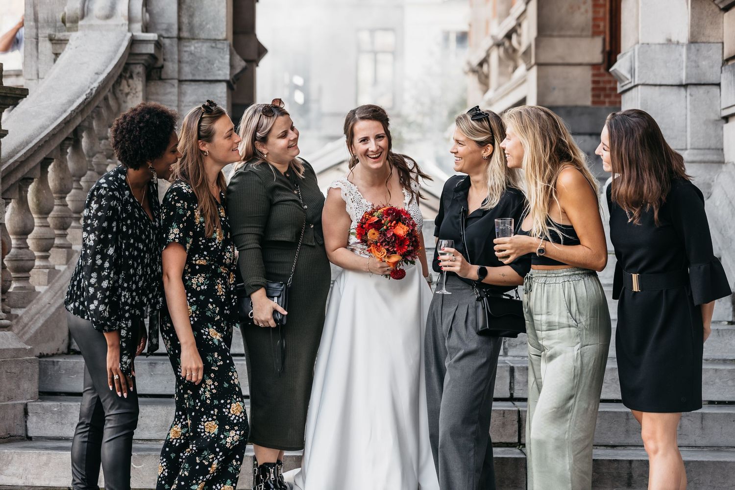 Bride laughing with bridesmaids on ornate stone steps holding a vibrant red bouquet.
