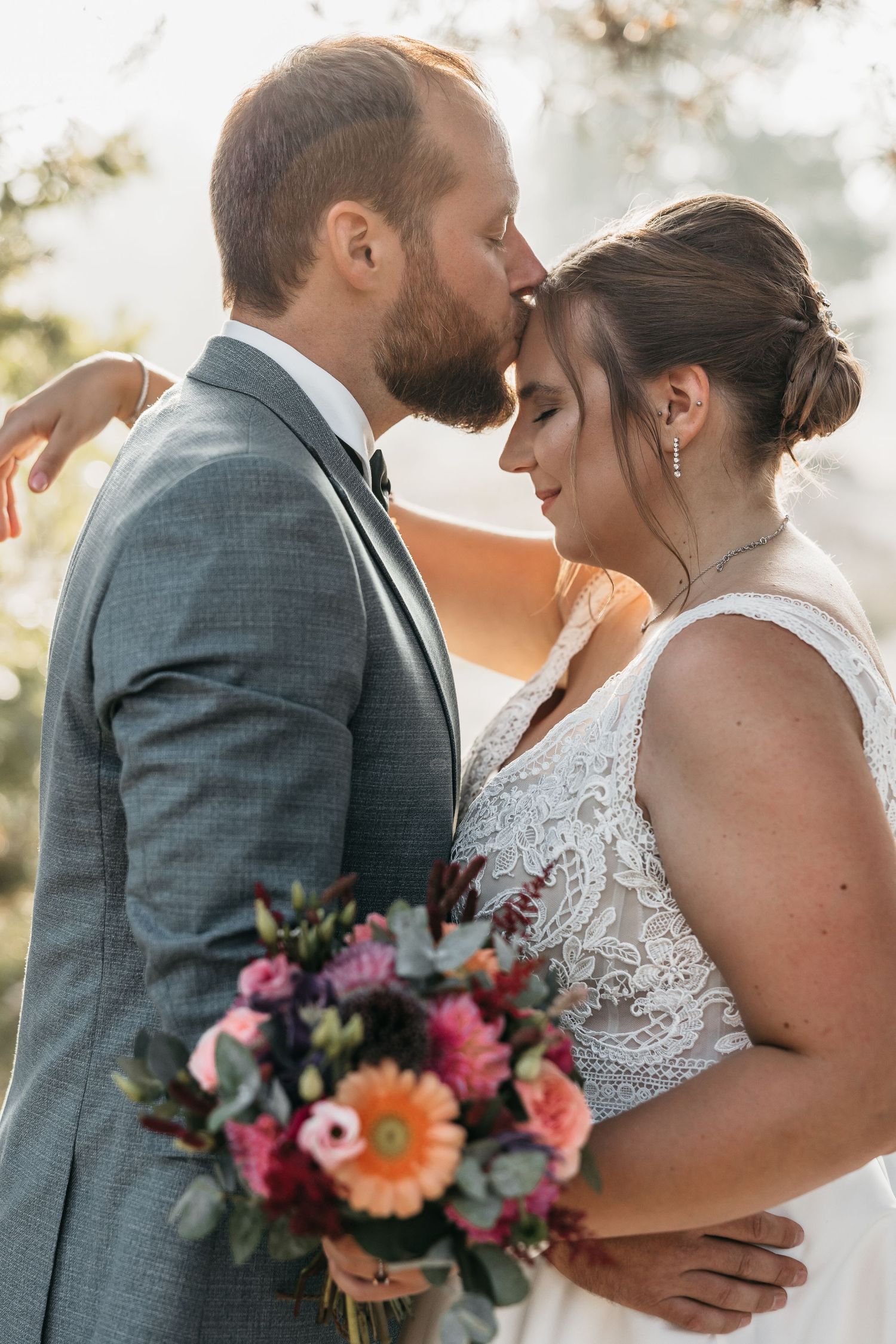 Groom kisses bride on forehead as she holds a vibrant colorful wedding bouquet in soft natural light.