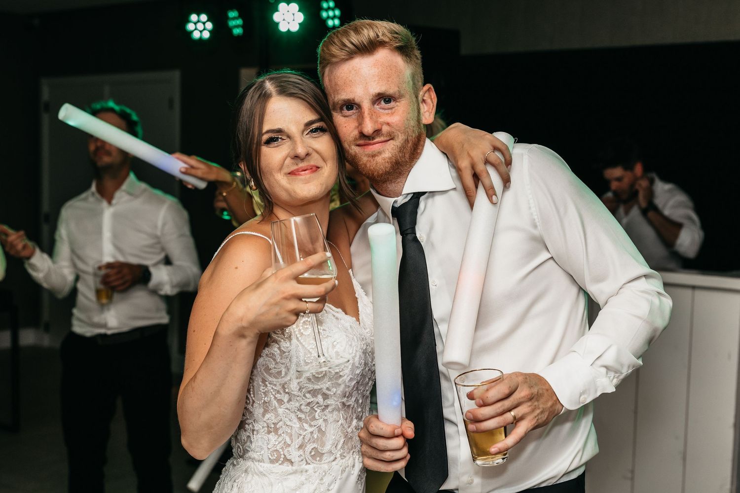 Smiling bride and groomsman pose with drinks and glow sticks at lively wedding reception.