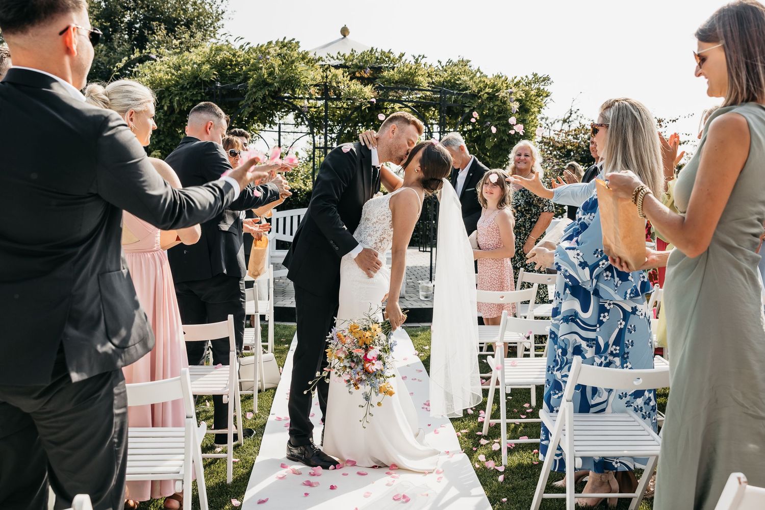 Bride and groom share a kiss on the aisle as wedding guests toss colorful flower petals.