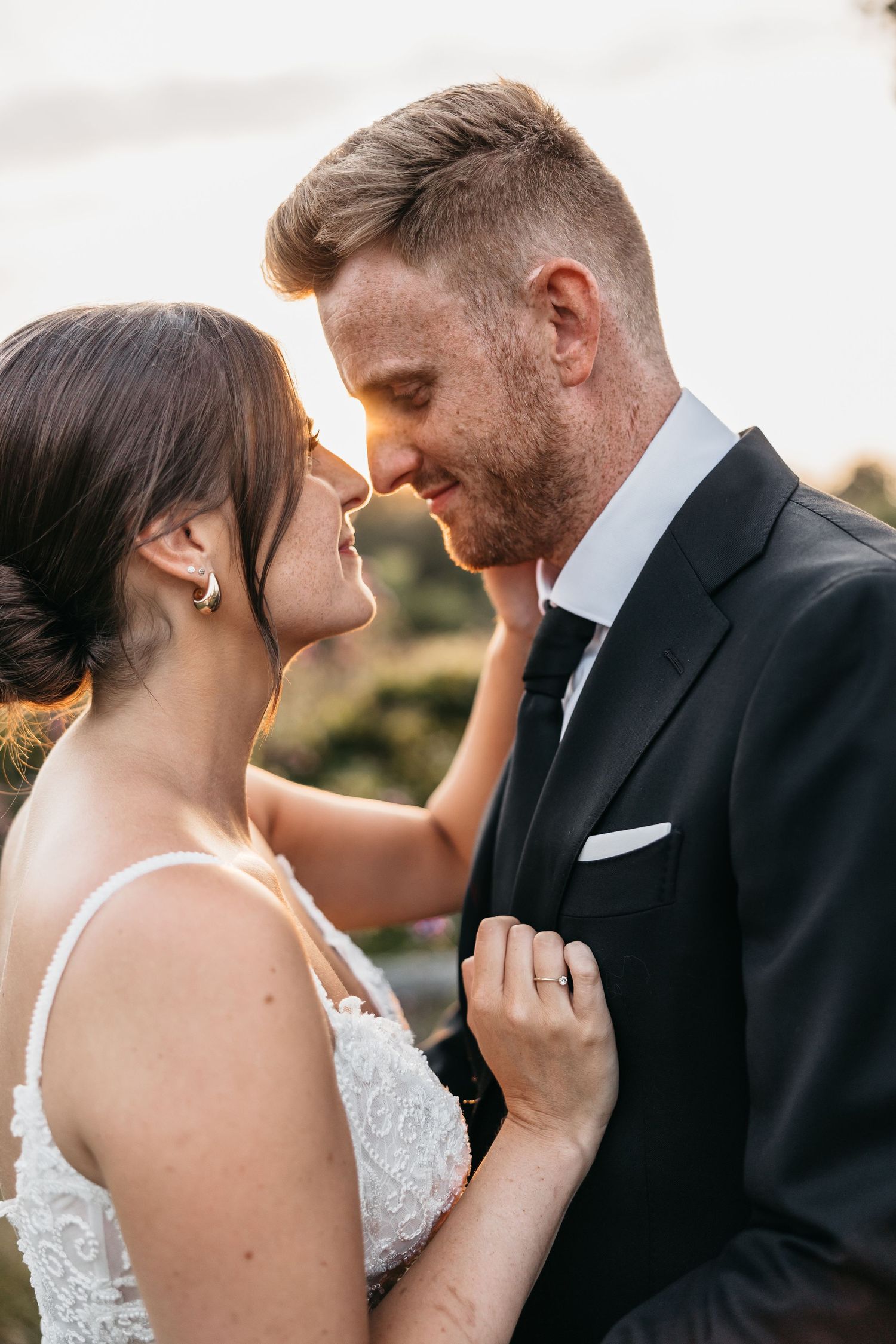 Smiling bride and groom lean in close together at sunset, bride in white lace dress gently touching groom's bearded face.
