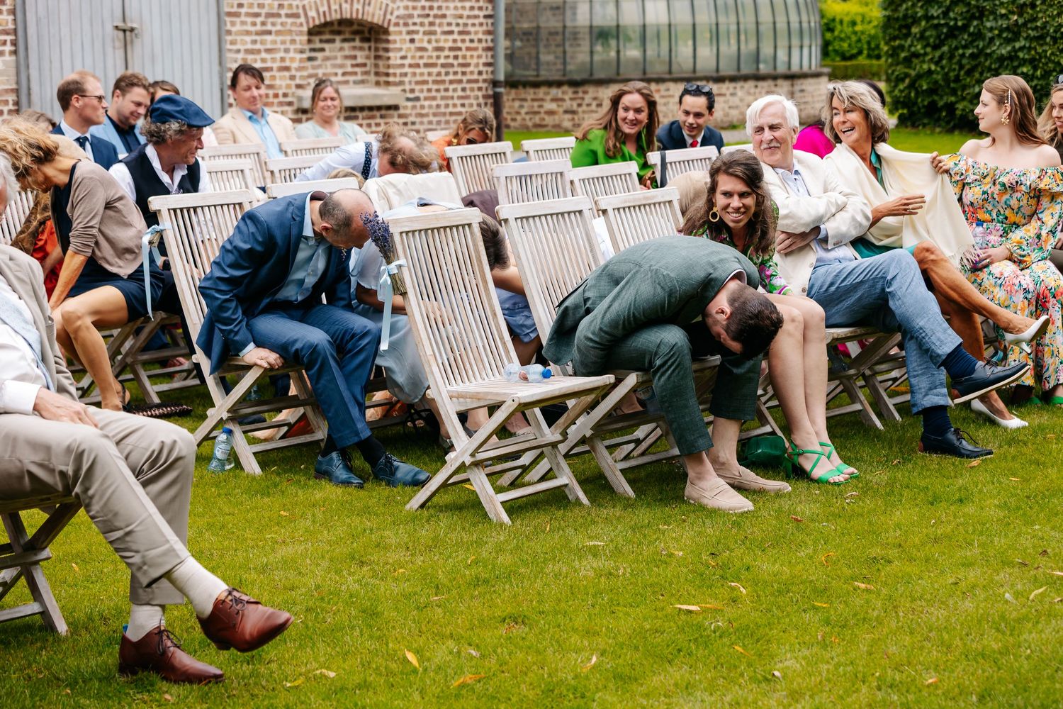 Wedding guests laughing and socializing on white garden chairs during outdoor ceremony reception on green lawn.