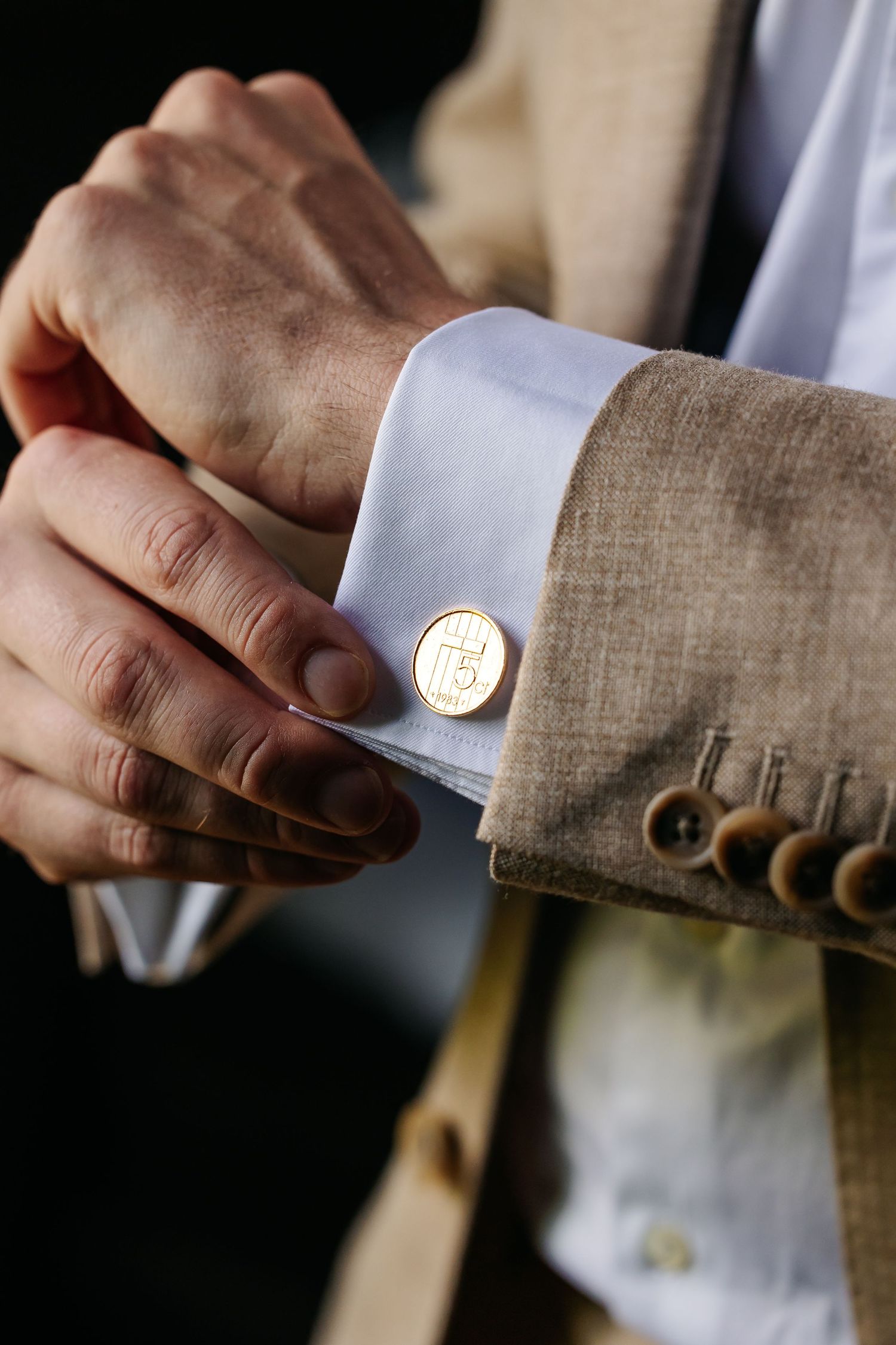 Groom fastening gold monogram cufflinks on white shirt with beige suit.