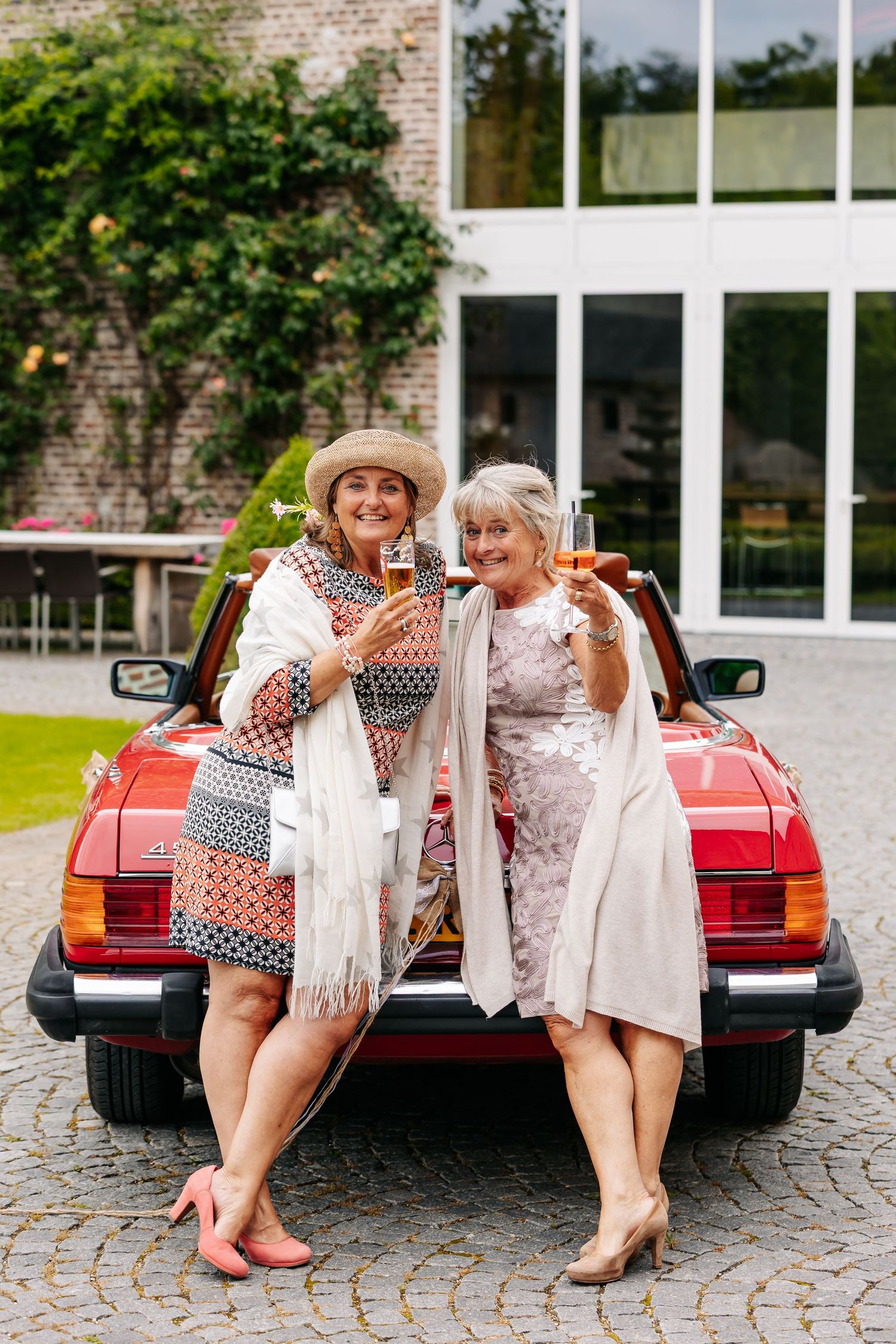 Two older women toasting champagne while leaning on red vintage convertible car at wedding.