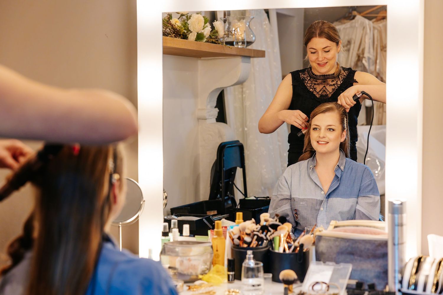 A hairstylist curls a smiling woman's hair in a vanity mirror salon setting with makeup tools on the counter.