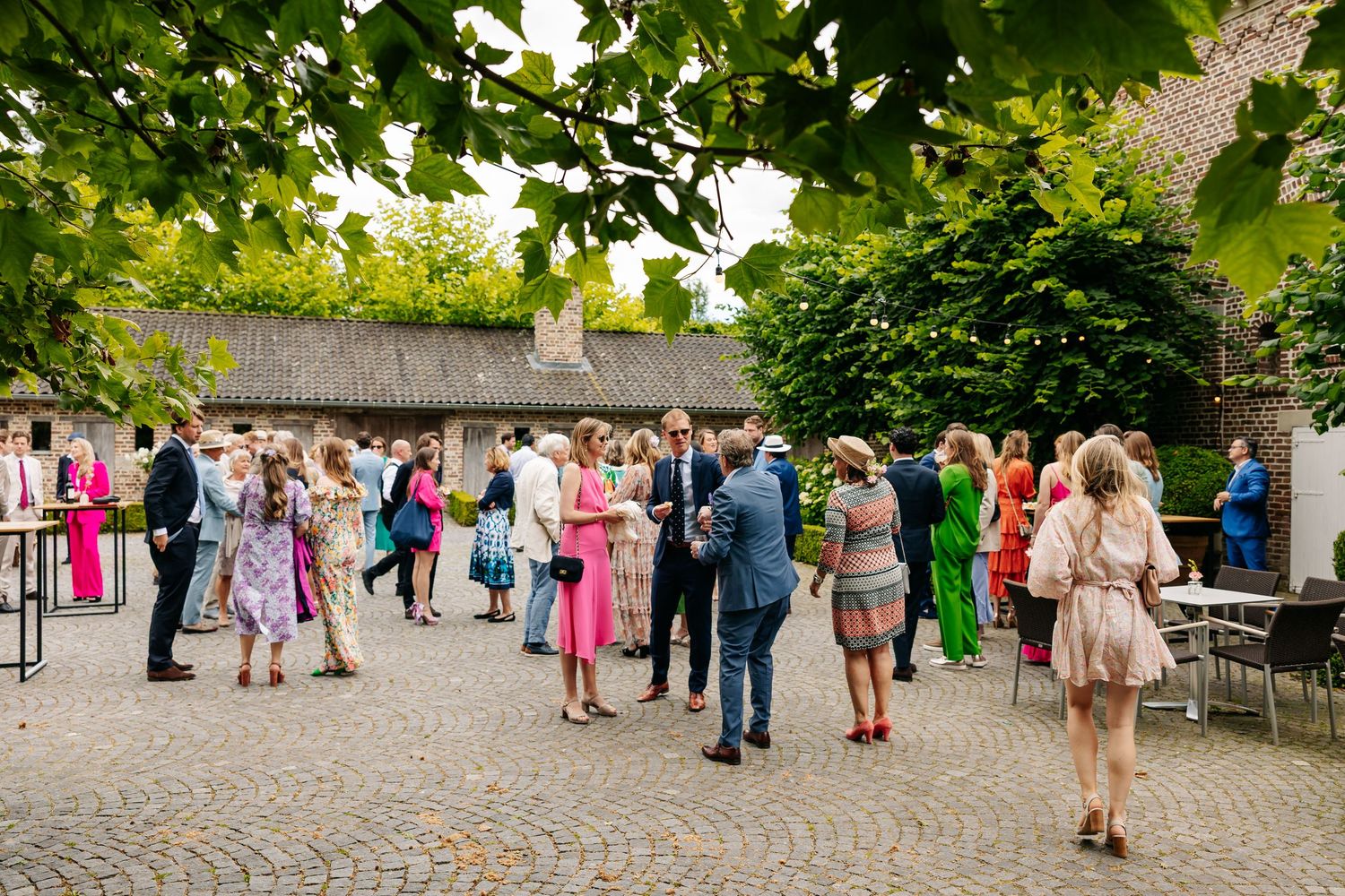 Elegantly dressed wedding guests socializing on a sunlit cobblestone courtyard surrounded by lush green trees.