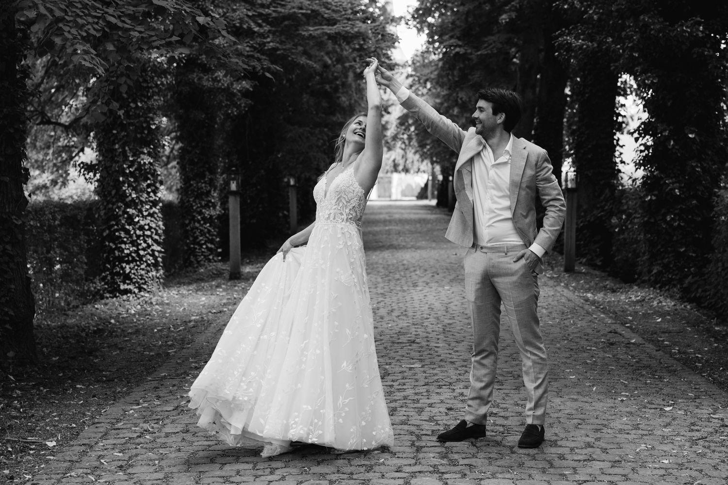 Black and white photo of bride and groom dancing joyfully on a tree-lined cobblestone path.