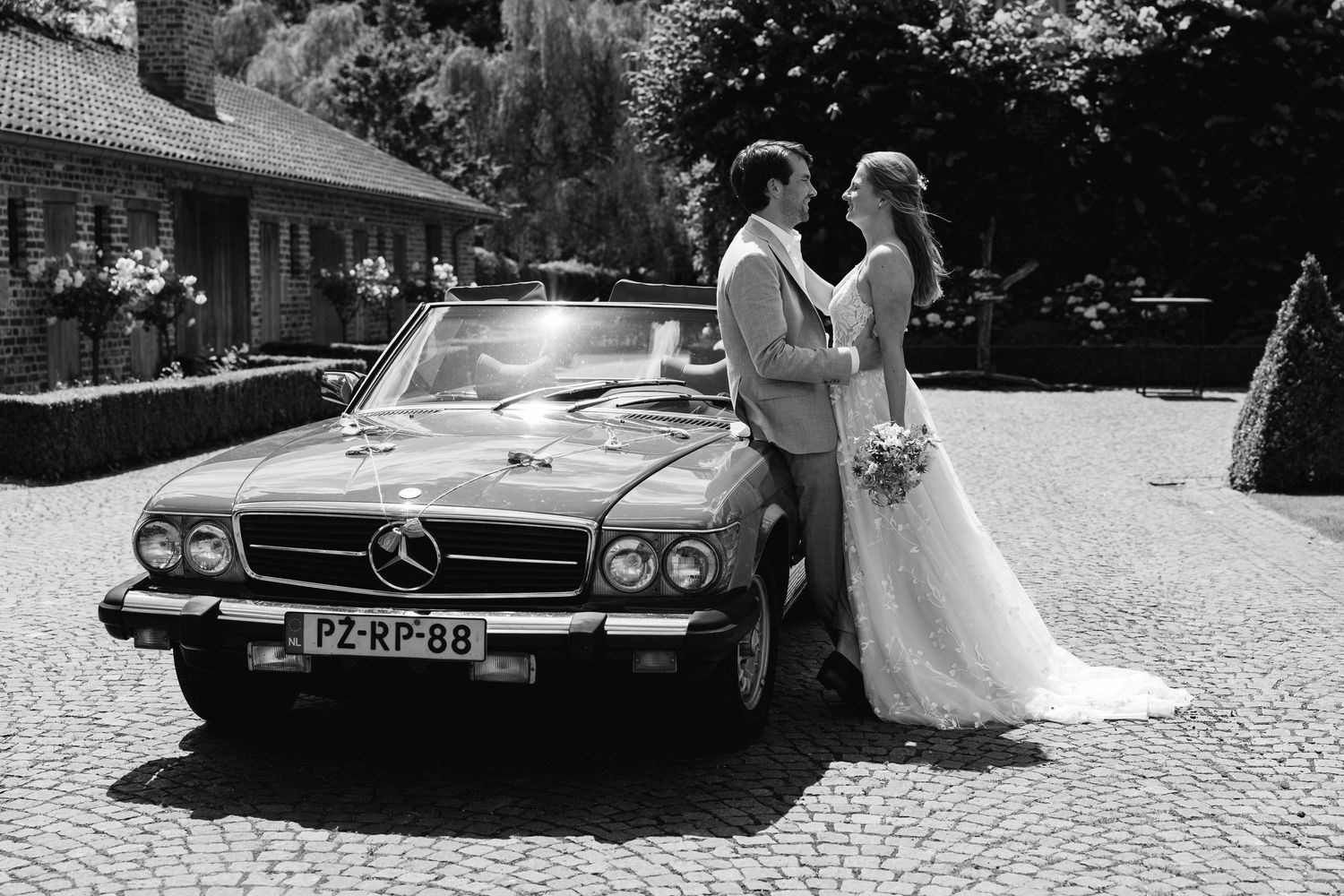 Bride and groom posing romantically beside vintage red Mercedes convertible in black and white.