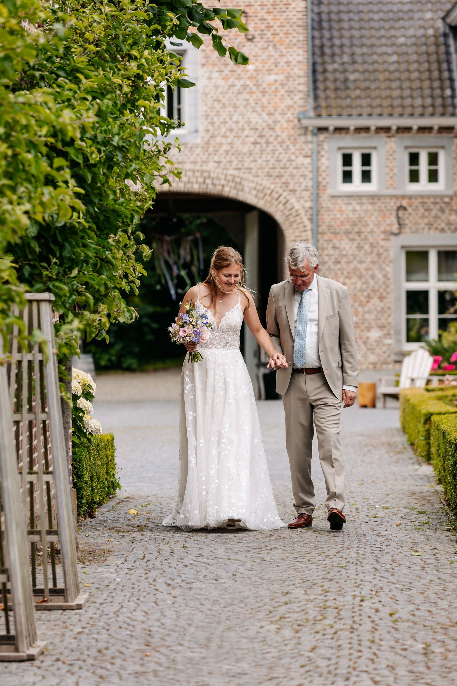 Bride in lace gown holding bouquet walks with older gentleman on cobblestone path at wedding venue.