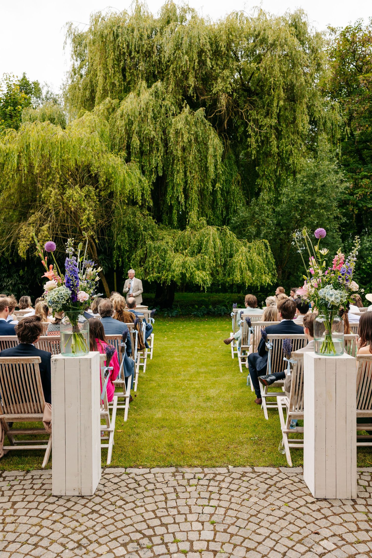Elegant outdoor wedding ceremony setup with floral arrangements, wooden chairs, and weeping willow backdrop.