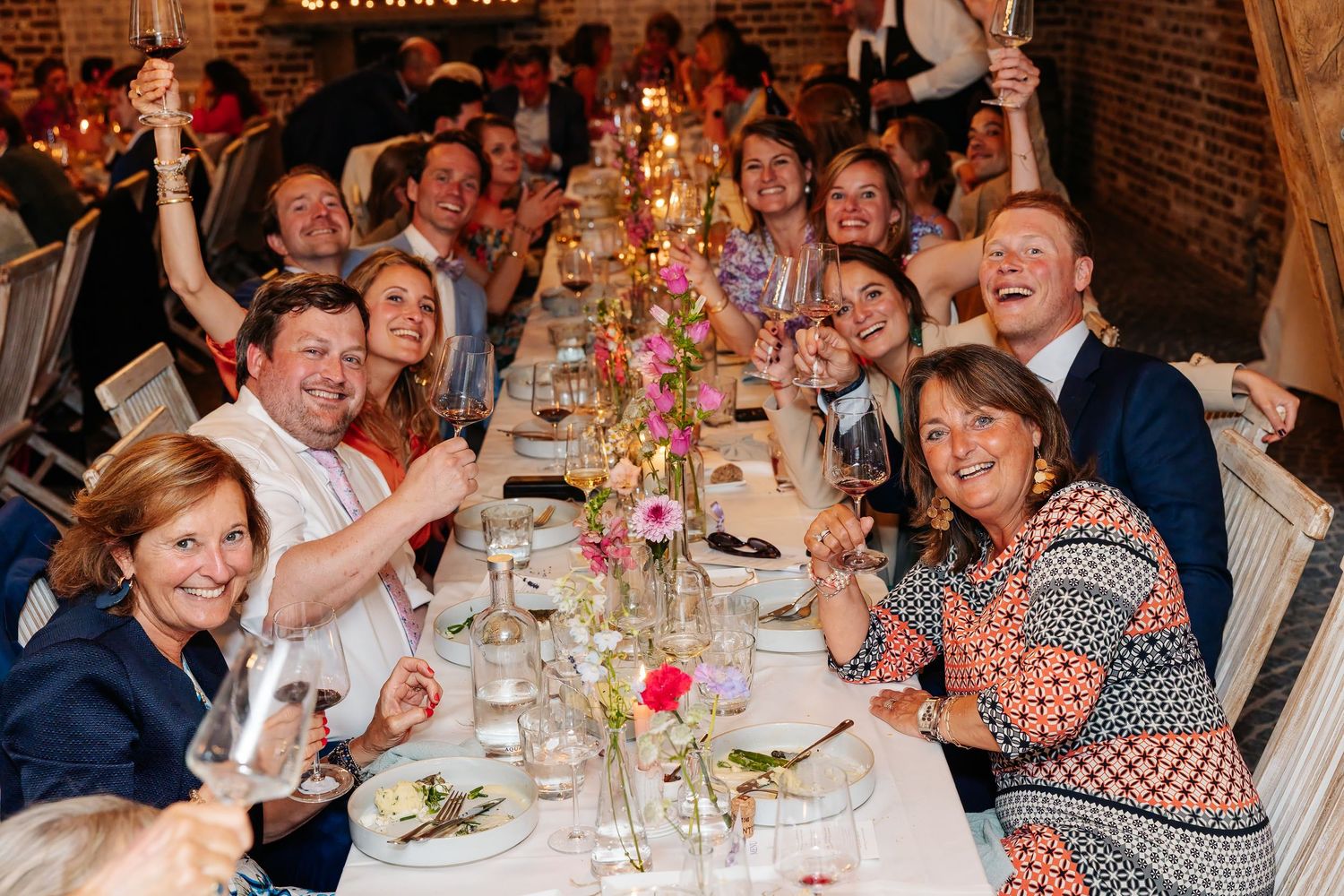 Joyful wedding guests raise glasses in a toast at a long decorated dinner table with floral centerpieces.