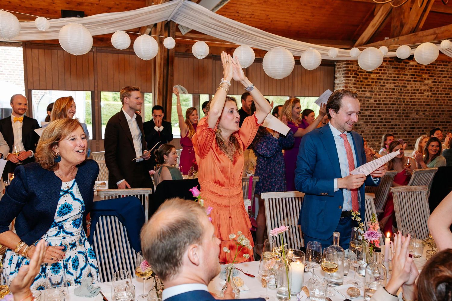 Joyful wedding guests celebrate at reception dinner in rustic venue decorated with white balloon ceiling.