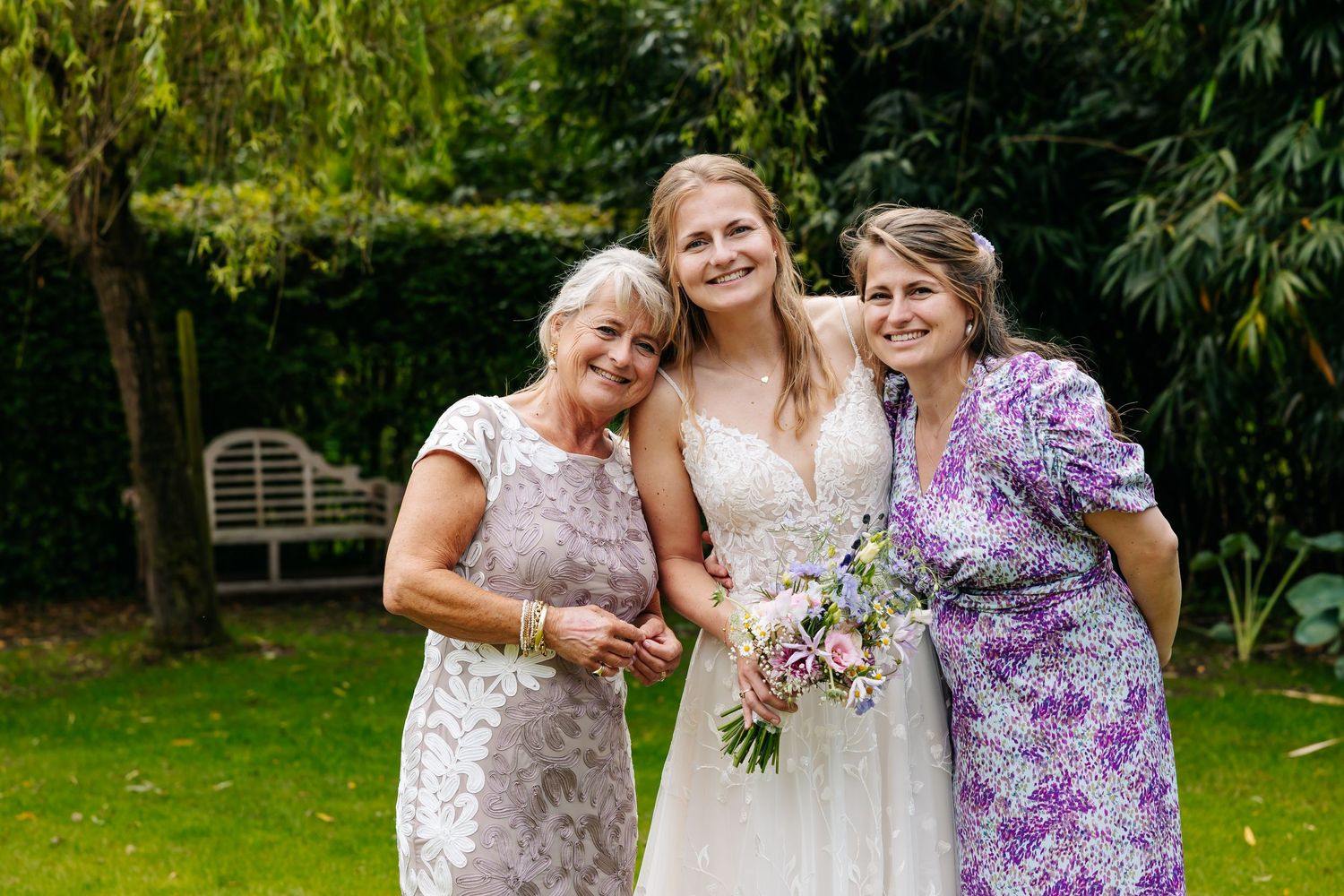 Bride in white lace gown poses with two smiling women in a lush garden setting.