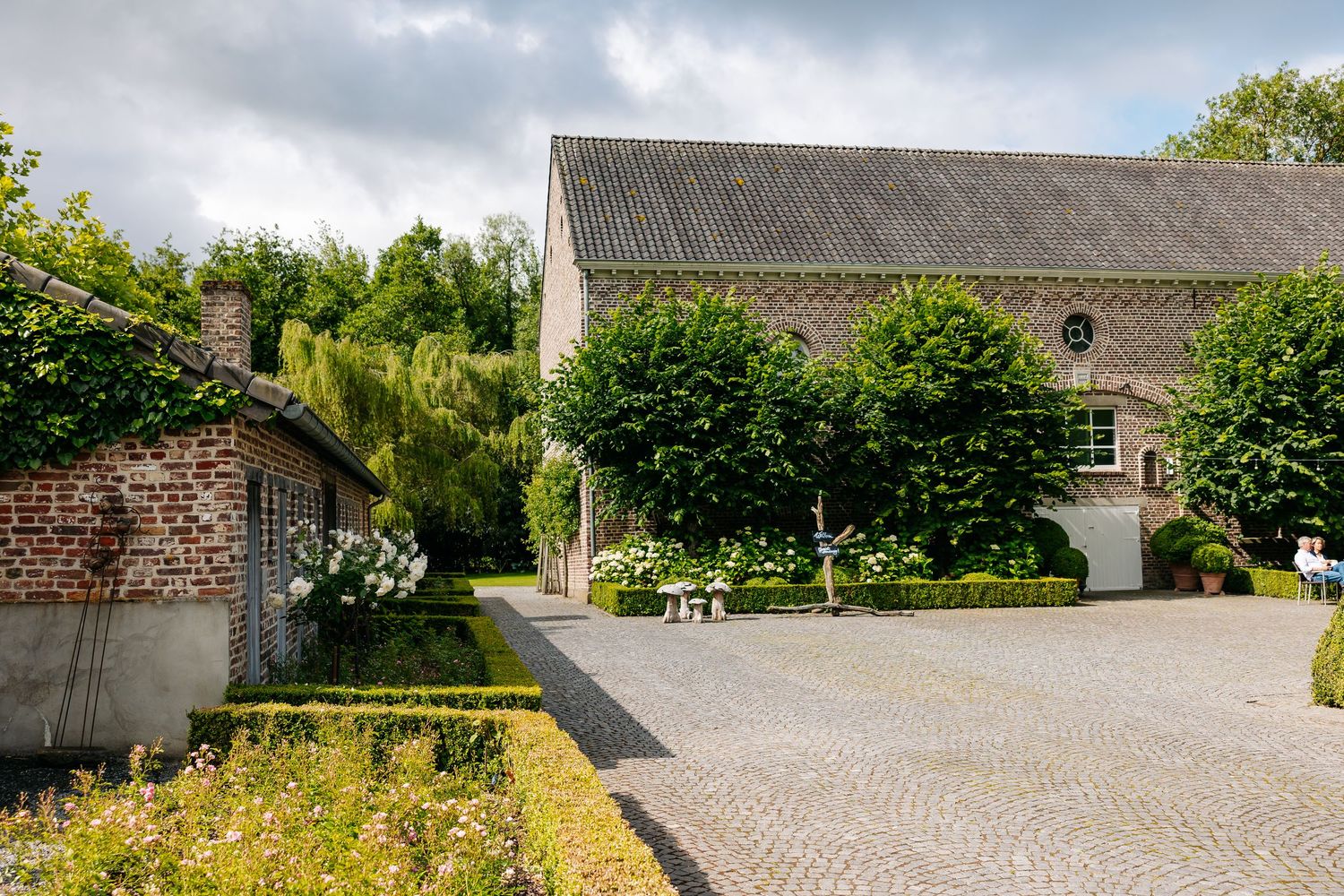 Charming Belgian farmhouse with cobblestone courtyard, manicured hedges, and lush green trees under a cloudy sky.
