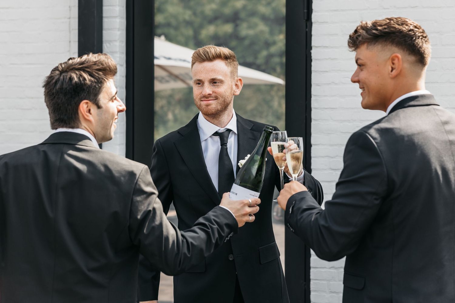 Three well-dressed groomsmen in black suits celebrate with champagne bottles and glasses.