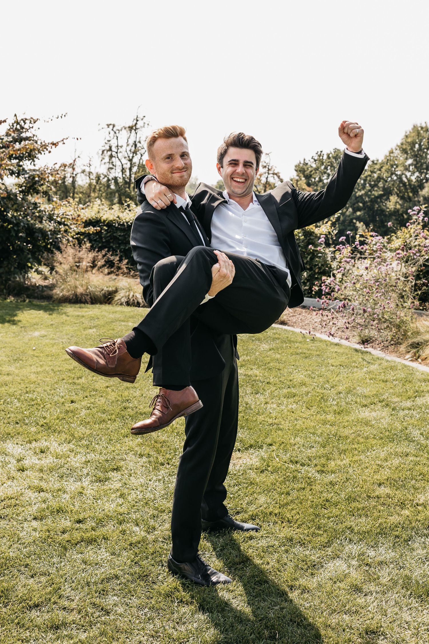 Two grooms celebrate joyfully outdoors, one lifting the other in a playful wedding photo.