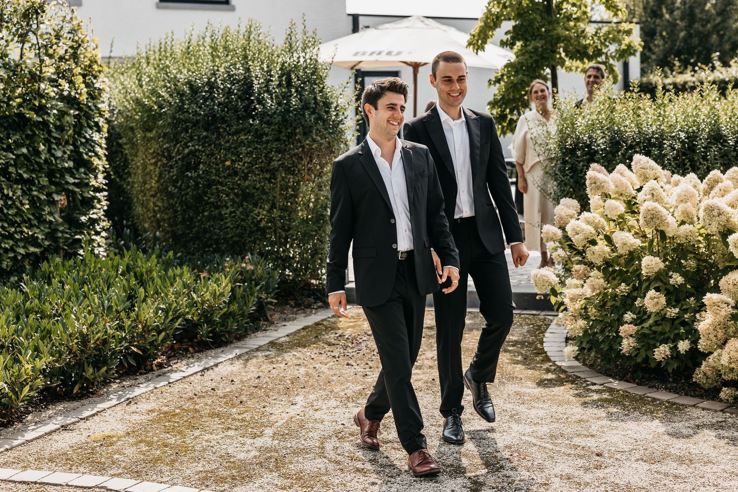 Two men in matching black suits walk together through a lush garden at a wedding venue.