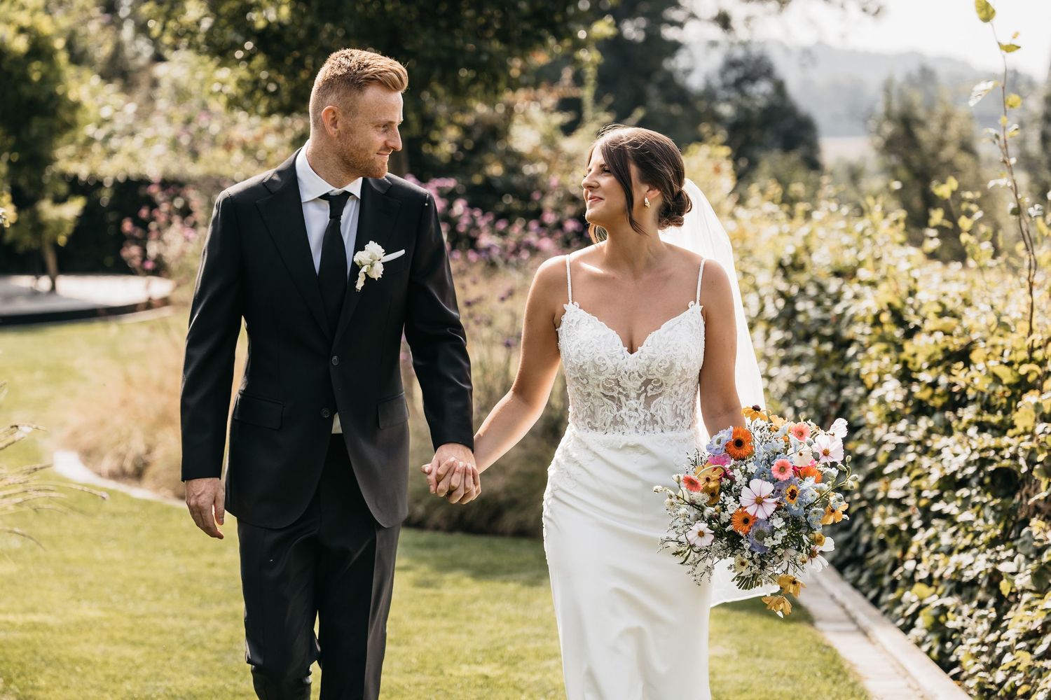 Happy bride and groom holding hands and smiling while walking through a sunny garden with colorful flowers.