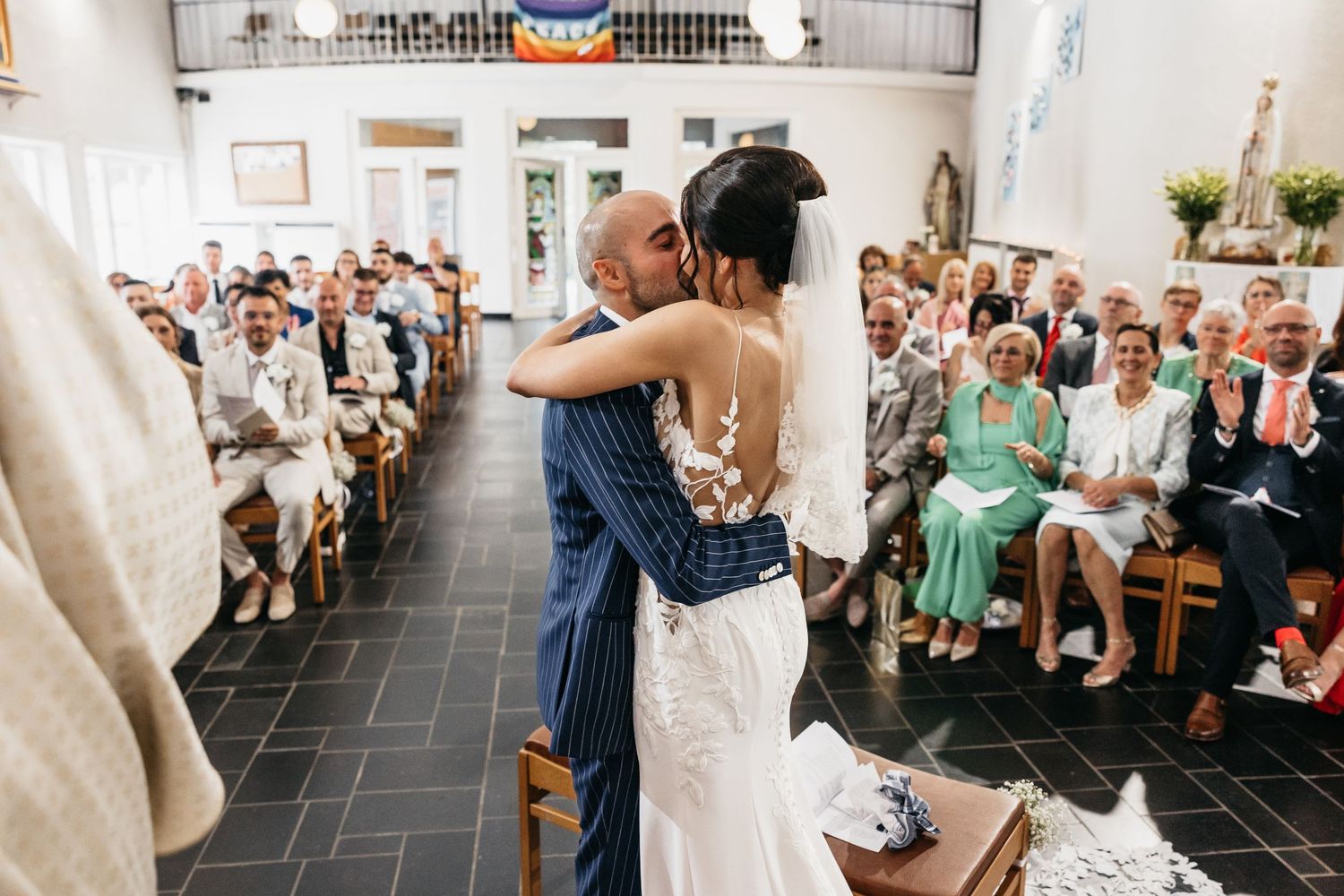 Newlywed couple sharing first kiss at altar with wedding guests seated in church pews.