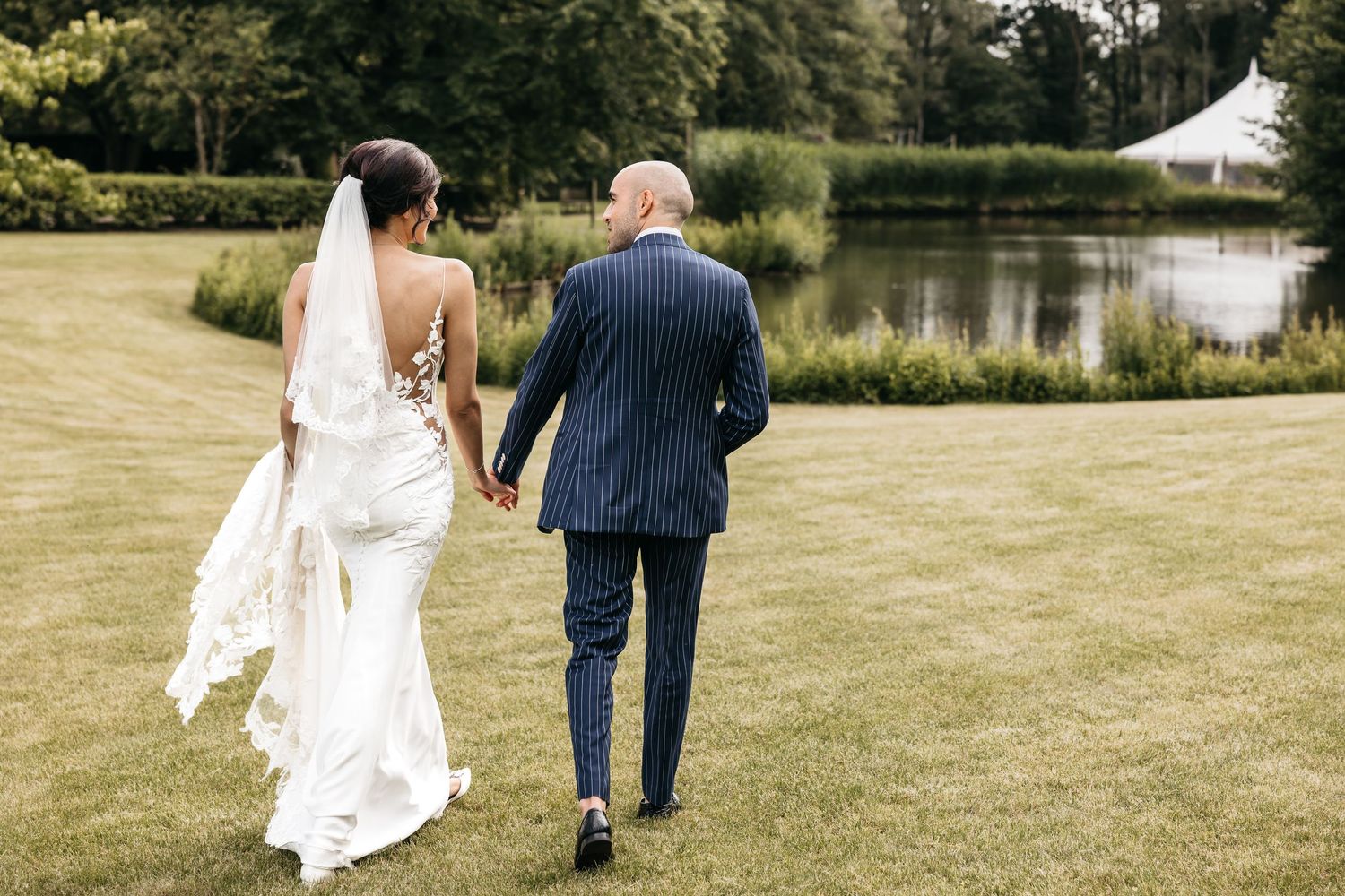 Bride and groom walking hand in hand across a green lawn toward a serene pond surrounded by trees.