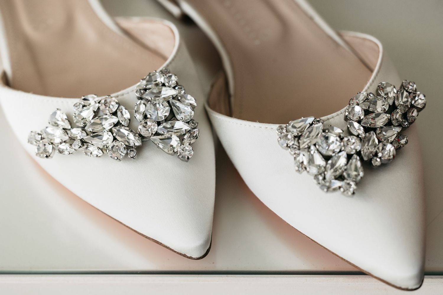 Close-up of white satin pointed toe bridal heels with sparkling crystal brooch detail.