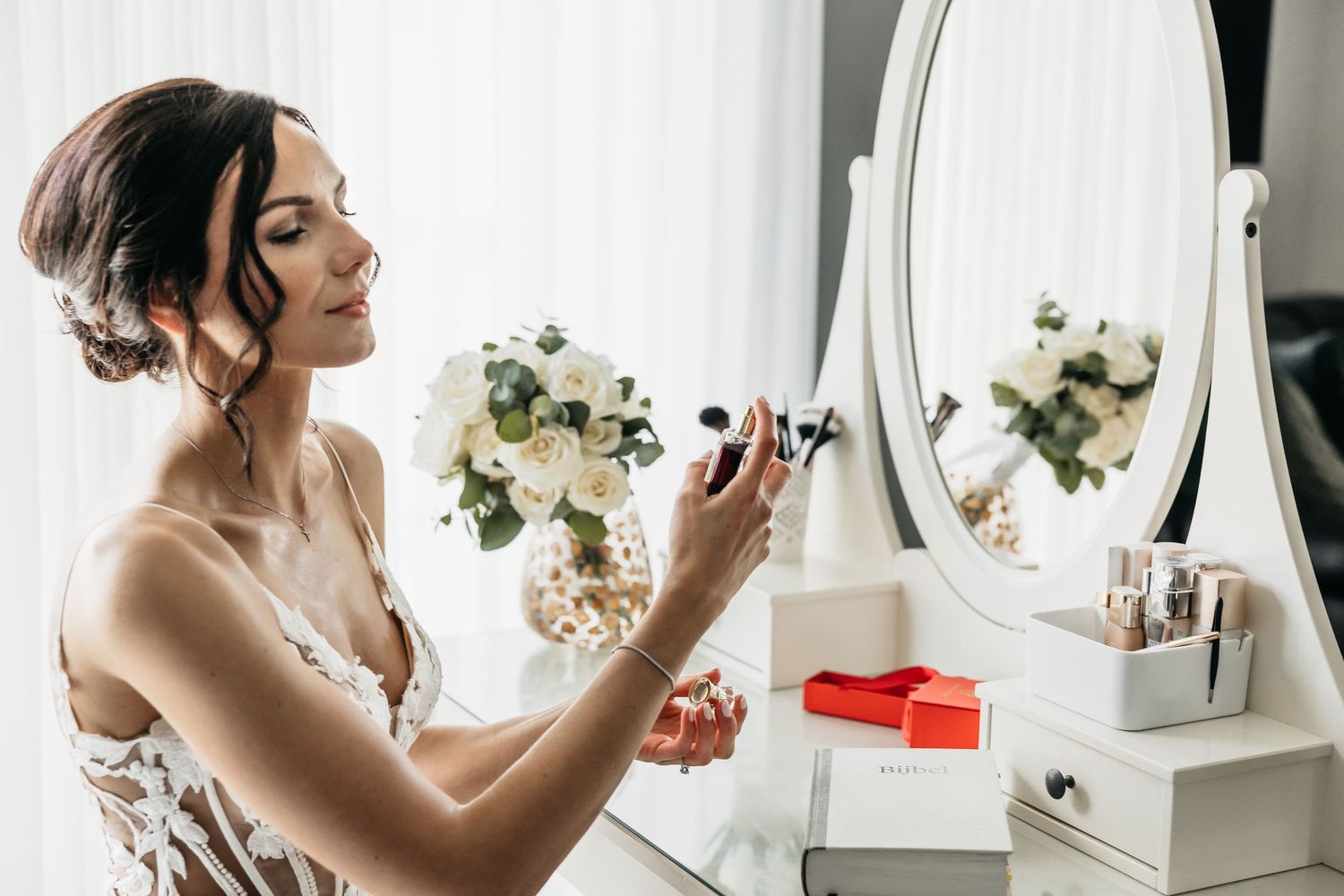 Elegant bride holding perfume bottle near a white vanity mirror with fresh white flowers on her wedding day.
