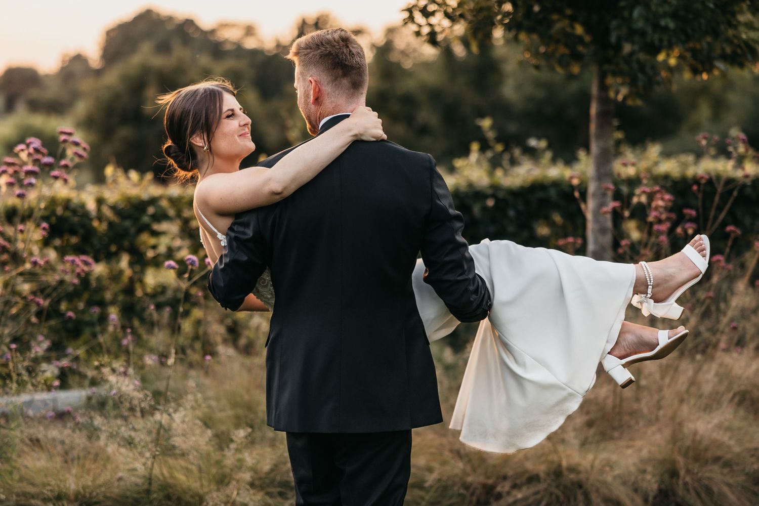 Groom in black suit carries bride in white wedding dress through a wildflower garden at warm sunset.